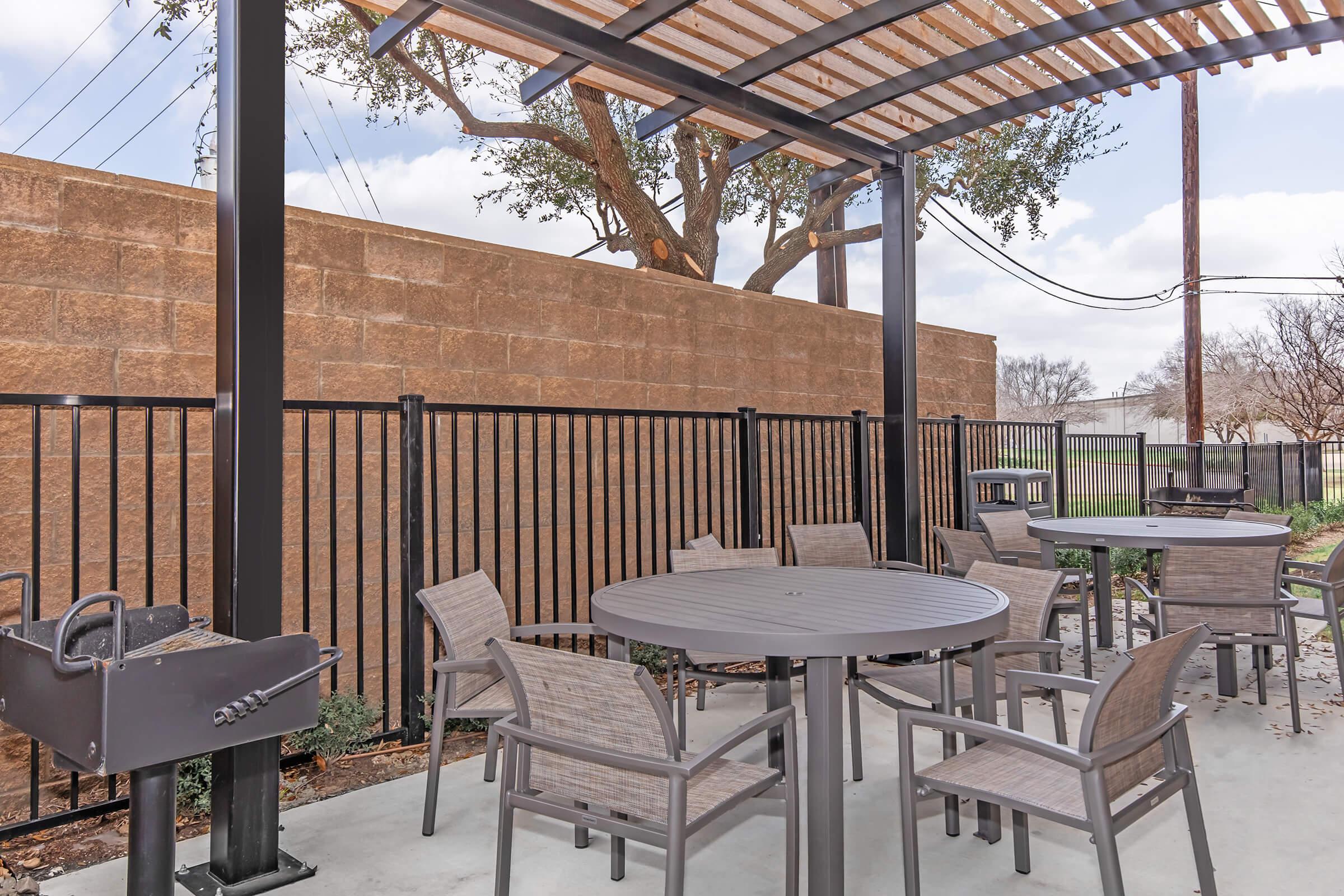 Outdoor seating area featuring several round tables and chairs under a shaded pergola. A grill is present nearby, with a backdrop of a tall, textured wall and some trees in the vicinity. The setting is well-maintained, ideal for gatherings or relaxation.