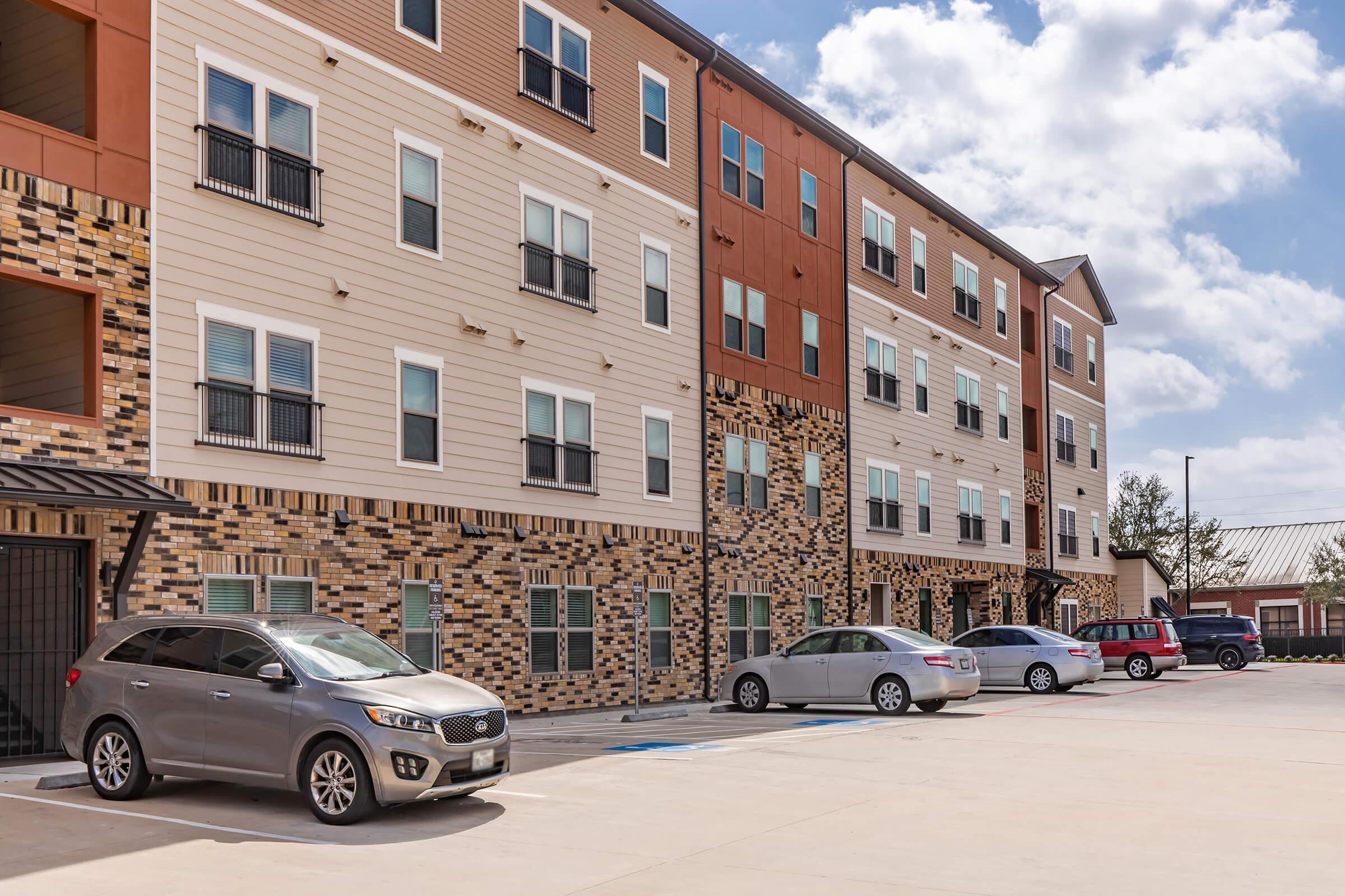 A multi-story residential building featuring a mix of brick and siding, with various windows and balconies. Several parked cars are visible in front of the building on a paved area, under a partly cloudy sky.