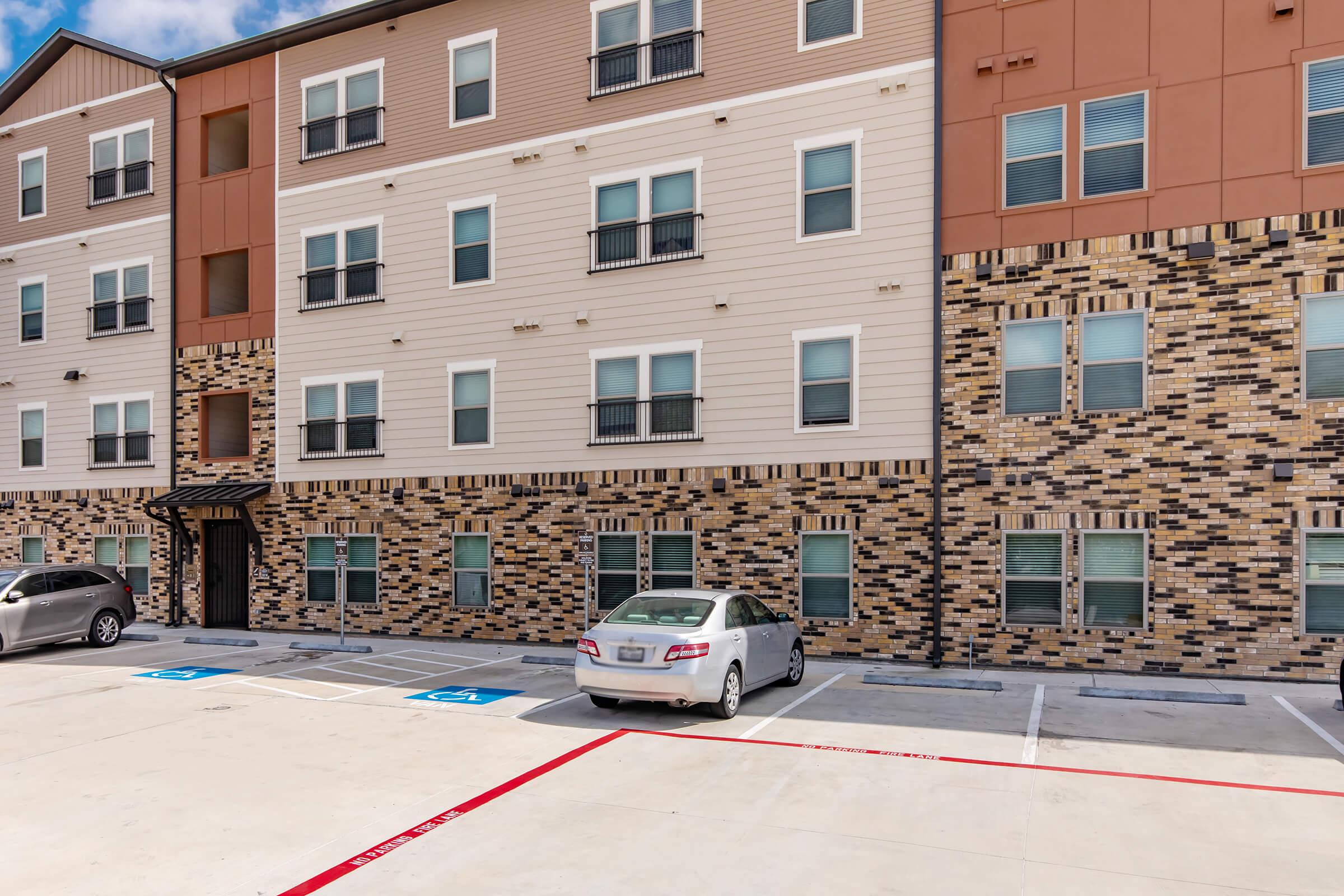 A modern apartment building with a multi-story exterior featuring a mix of tan and dark brick. There are several windows with black railings, and a few parking spaces in front, including a designated accessible parking spot. The sky is bright and partly cloudy.