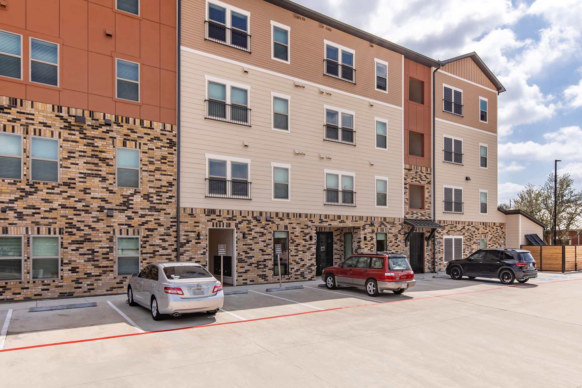Exterior view of a modern apartment building featuring a combination of brick and siding materials. The building has multiple floors with several windows. In front, there are several parked cars on an asphalt driveway, and cloudy skies are visible above.