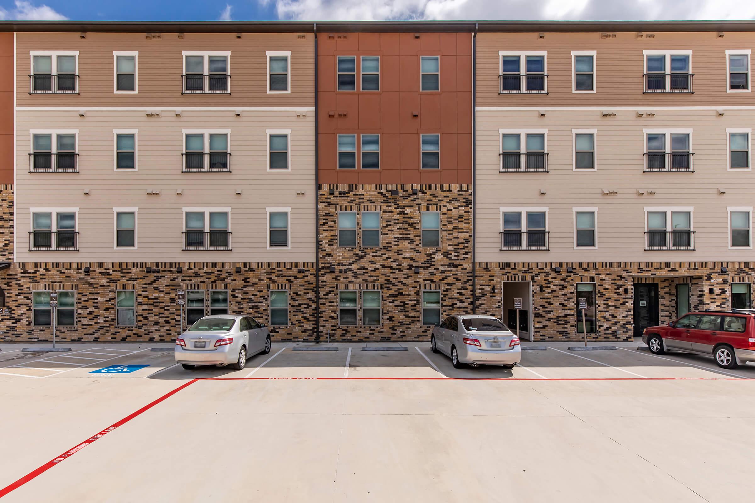 A modern apartment building featuring multiple stories with a combination of brick and siding. The facade includes various window designs, and there is a parking area in front with several parked cars, including a red vehicle. The sky is partly cloudy, adding a touch of contrast to the structure.