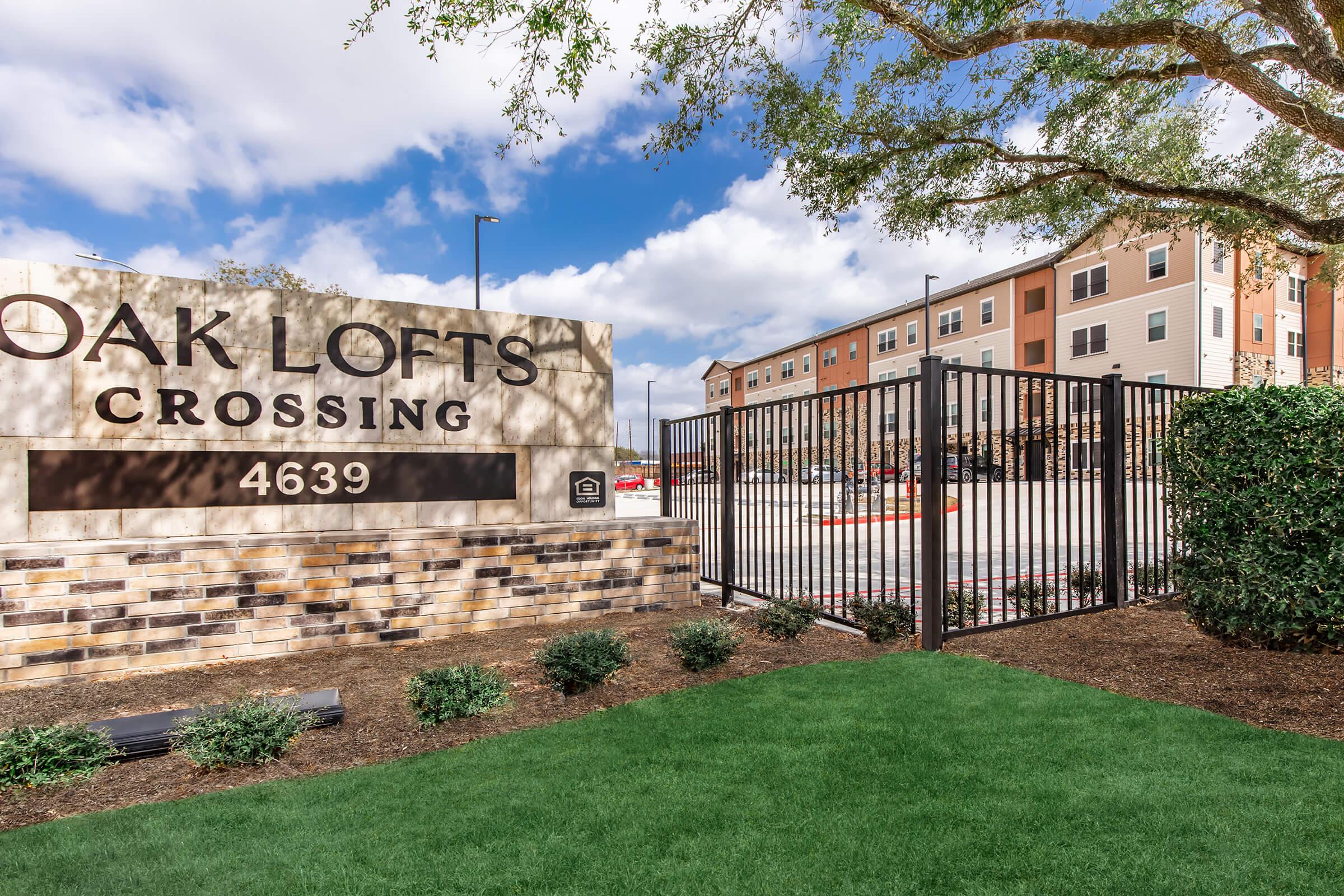 Sign for "Oak Lofts Crossing" featuring the address 4639, surrounded by greenery and a gated entrance. In the background, an apartment building is visible under a partly cloudy sky. The scene conveys a welcoming atmosphere with a well-maintained exterior.
