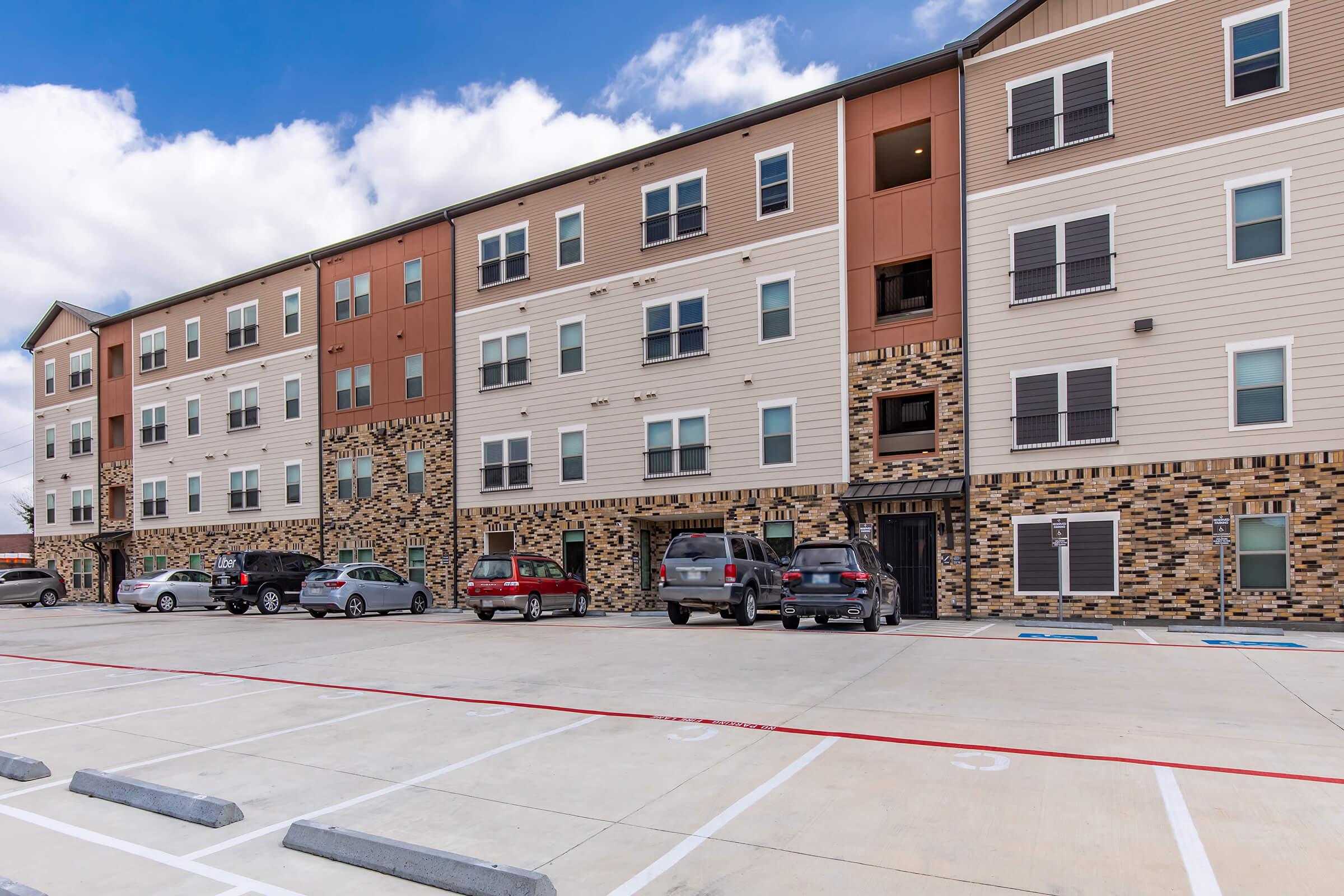 A modern multi-story apartment building featuring a mix of brown and beige cladding. The facade includes decorative stonework, numerous windows, and outdoor parking with multiple parked vehicles. The sky is partly cloudy, adding to the contemporary urban setting.