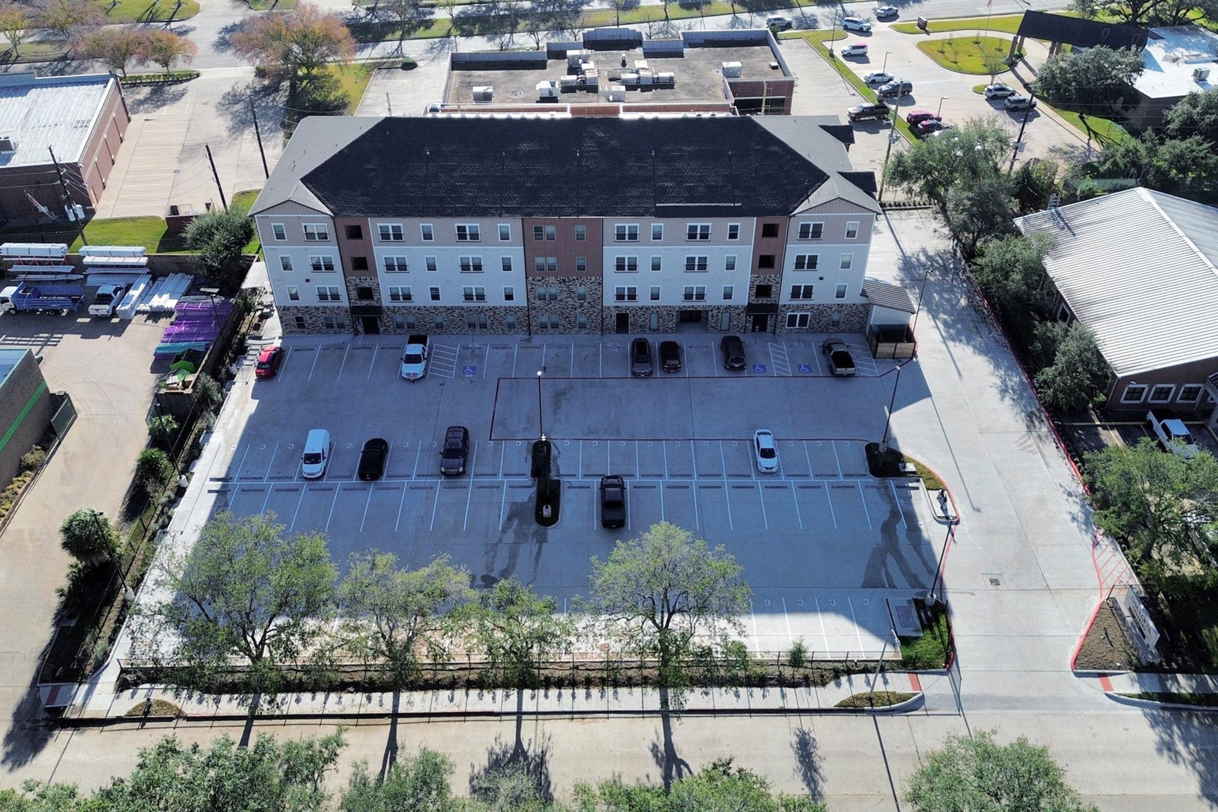 Aerial view of a multi-story building with a parking lot. The building features a mix of brick and siding, surrounded by small trees. The parking lot shows several parked cars and marked spaces. Nearby structures and landscaping are visible, with a clear sky in the background.