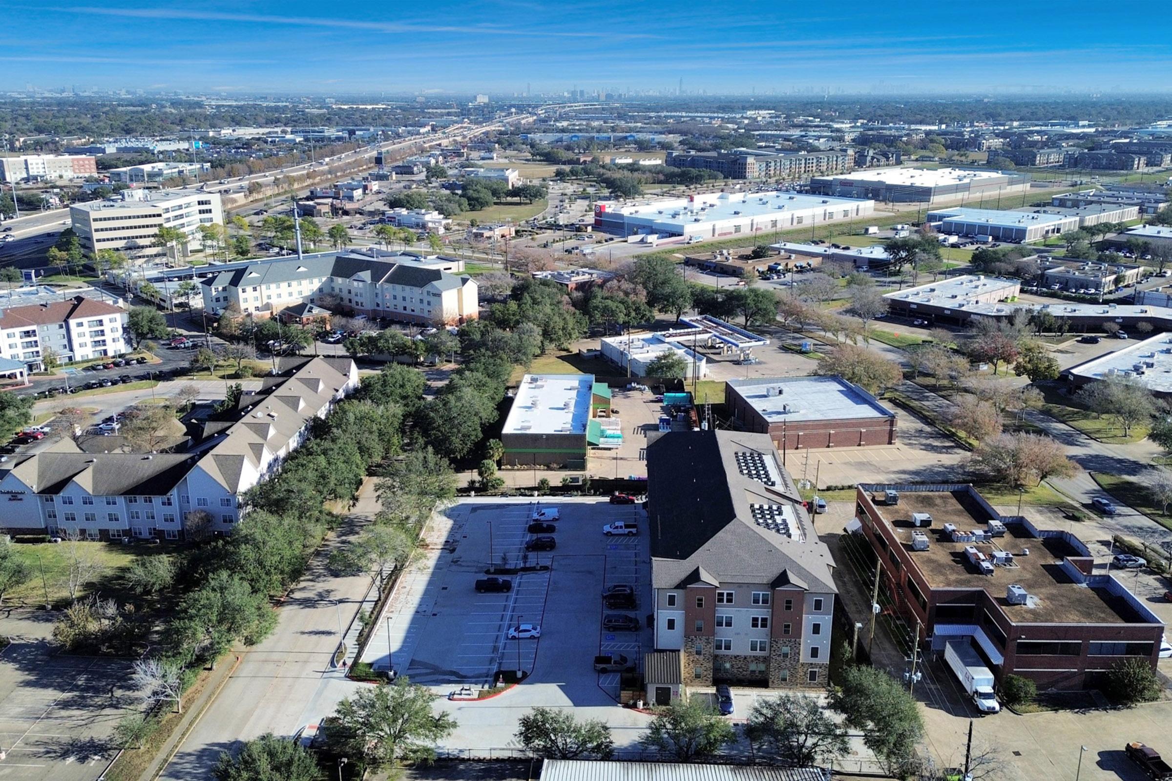 Aerial view of an urban landscape featuring residential buildings, commercial establishments, and a highway. The foreground includes a parking lot and green trees, while the background shows a city skyline in the distance under a clear blue sky.