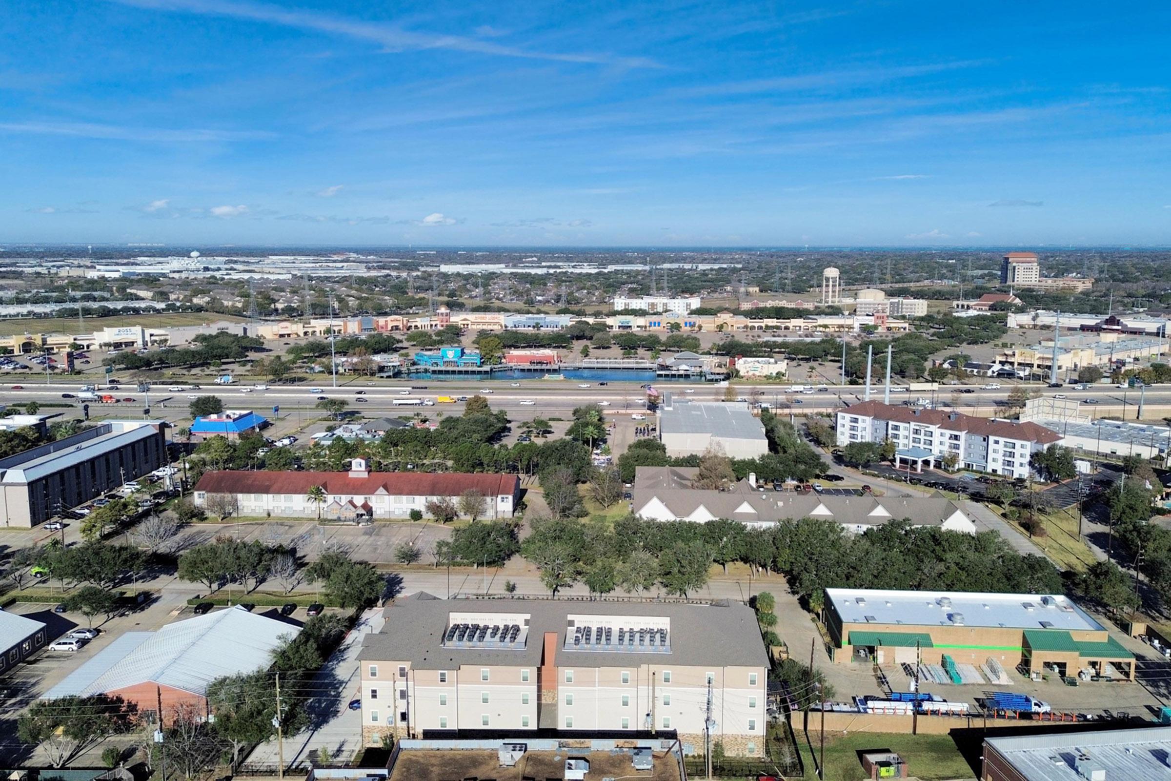 Aerial view of a city landscape featuring a mix of residential and commercial buildings, parking lots, and green spaces. In the background, highways and industrial areas are visible, with scattered trees and a clear blue sky overhead.