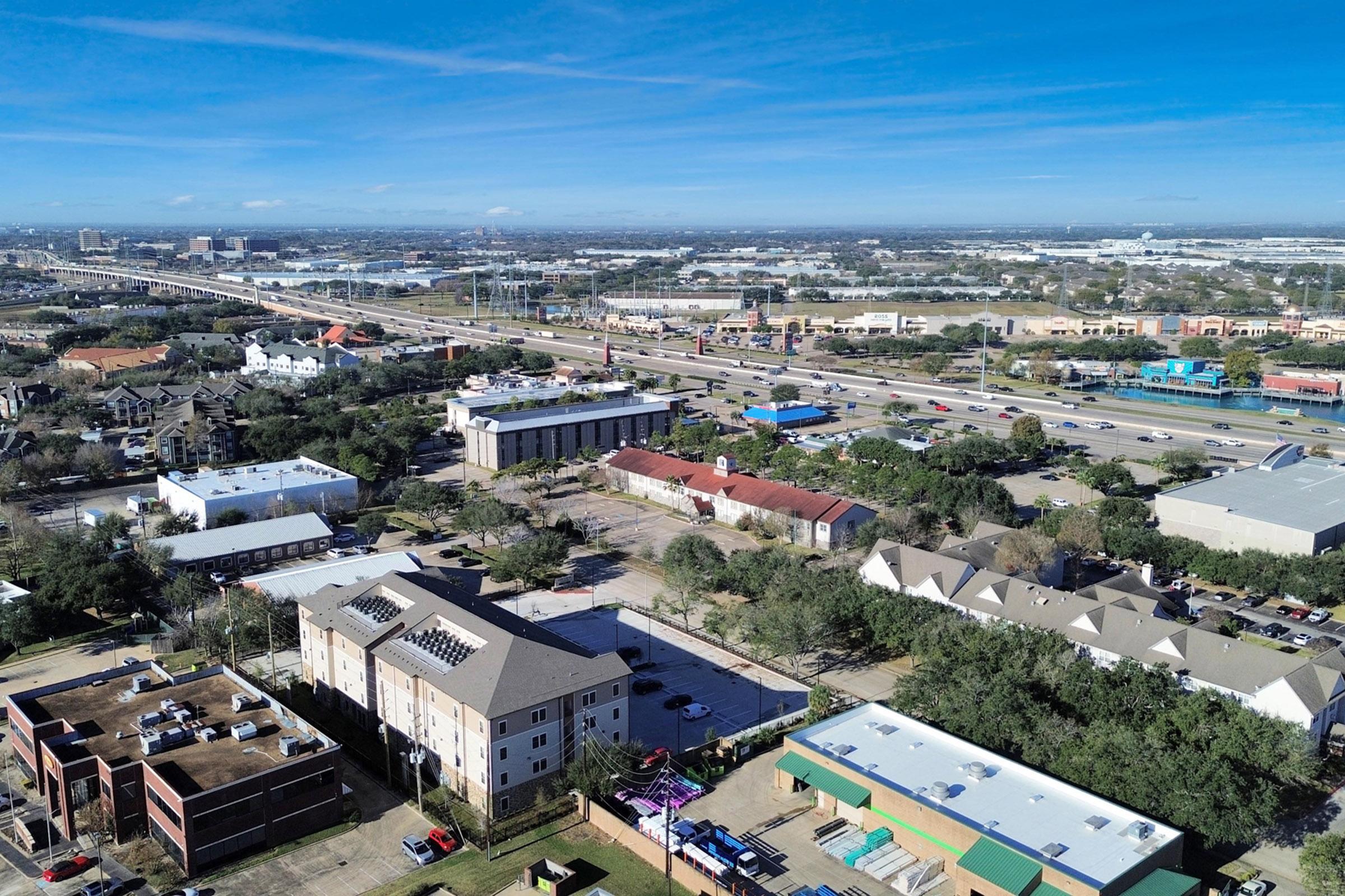 Aerial view of a suburban area featuring a mix of residential and commercial buildings, roads, and green spaces. Highways and vehicles are visible, along with a skyline in the distance under a clear blue sky. The image captures the layout and structure of the neighborhood with various architectural styles.