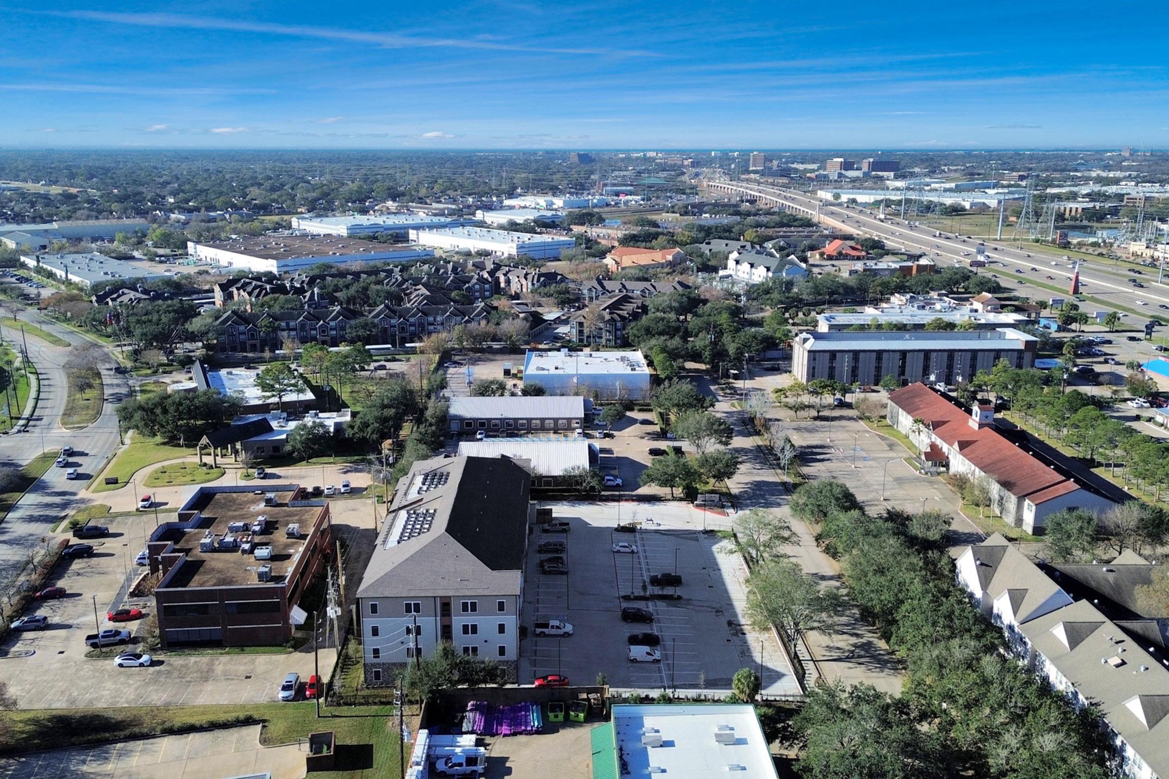 Aerial view of a suburban area featuring a mix of residential and commercial buildings. Green trees dot the landscape, with a clear blue sky above. In the background, highways and larger commercial lots are visible, indicating a bustling urban environment in the distance.