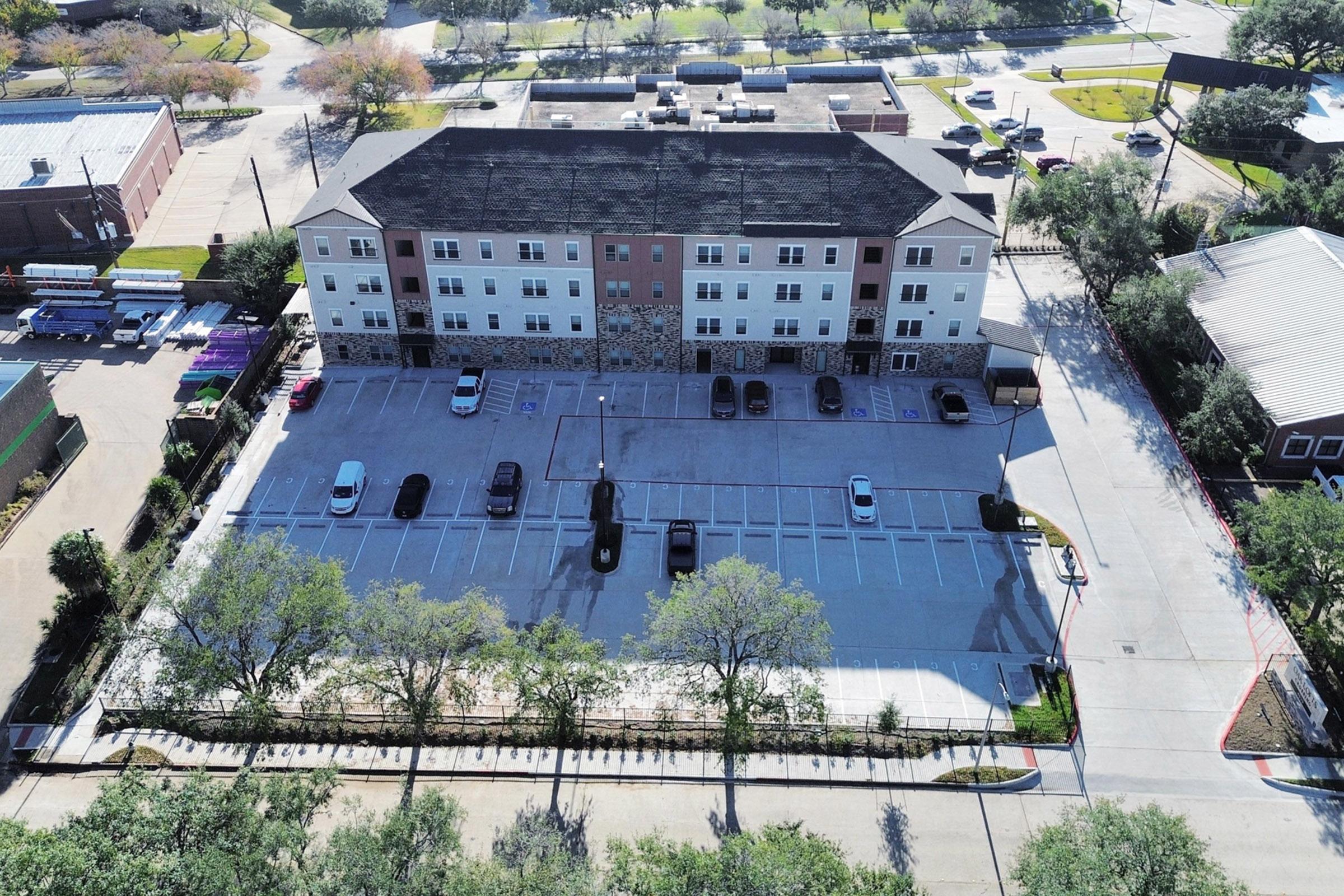 Aerial view of a multi-story building with a mix of brick and stucco exterior, surrounded by a parking lot filled with vehicles. Lush greenery lines the edges, with paved walking paths visible. The setting appears to be in a suburban area, with additional buildings and roads in the background.