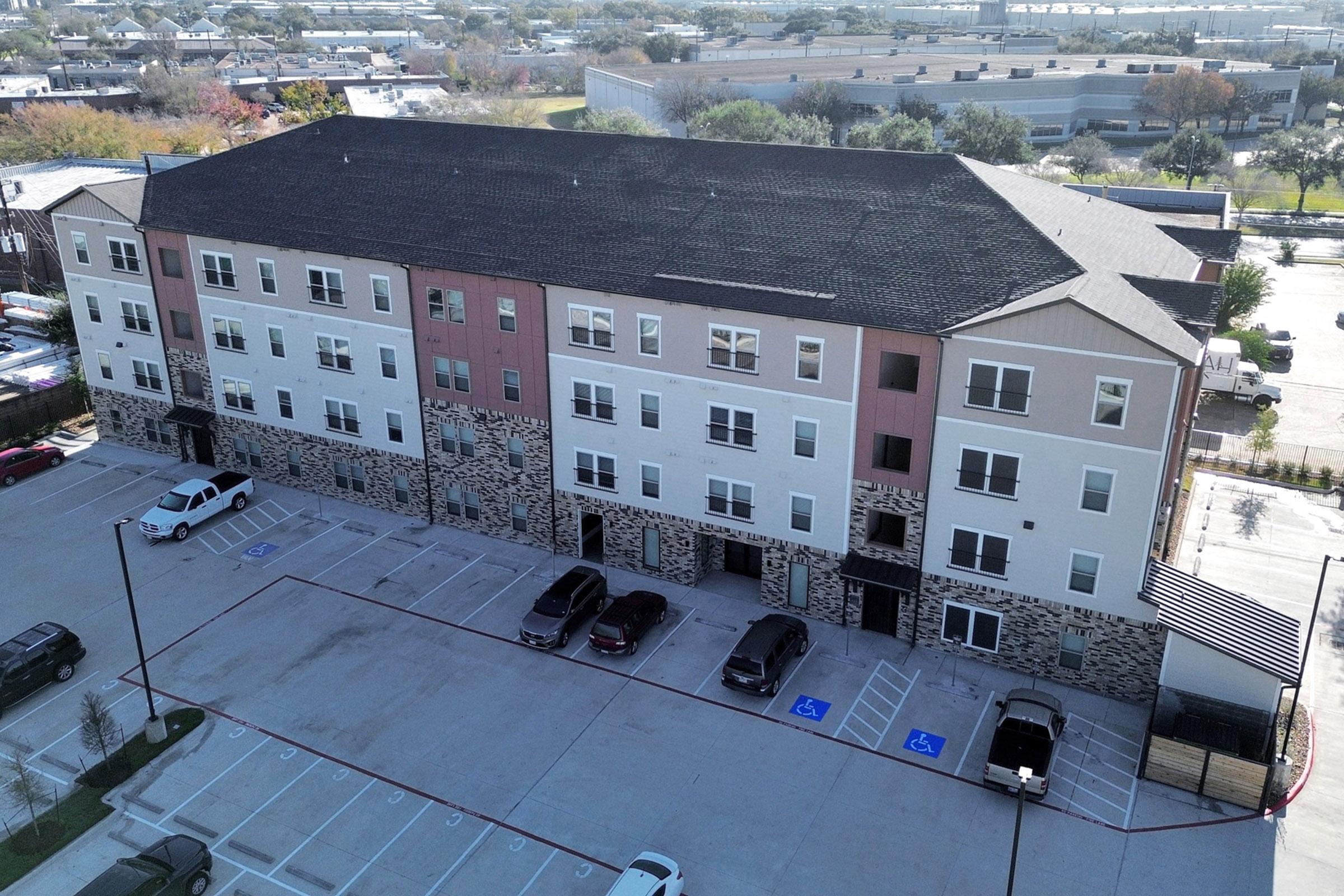A multi-story residential building featuring a mix of beige and pink exterior colors. The building has a stone facade at the base and multiple windows. A parking lot is in the foreground with several parked cars, including designated handicapped spaces, and trees in the background.