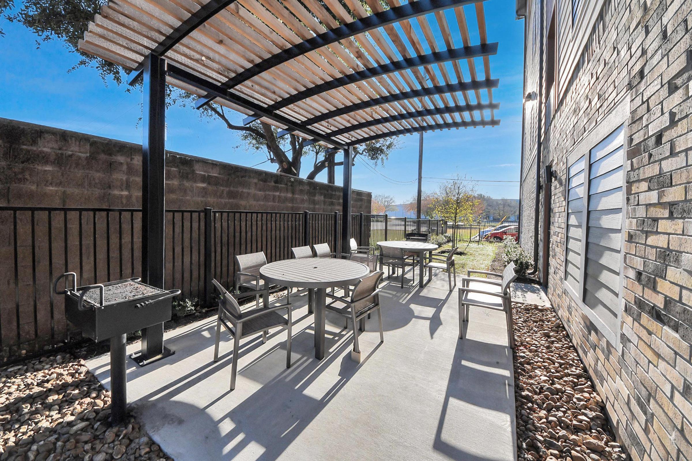 Outdoor patio area featuring a shaded canopy, tables, and chairs surrounded by a stone border. A small grill is placed nearby, and a clear blue sky is visible overhead. The space offers a relaxed atmosphere for gatherings.