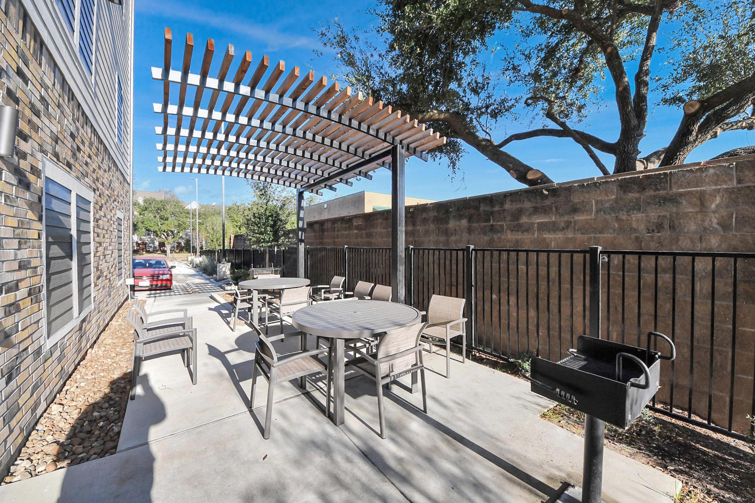 Outdoor patio area featuring a pergola, several round tables with chairs, and a charcoal grill. The setting is surrounded by greenery and a stone wall, under a clear blue sky. Ideal for gatherings and relaxation.