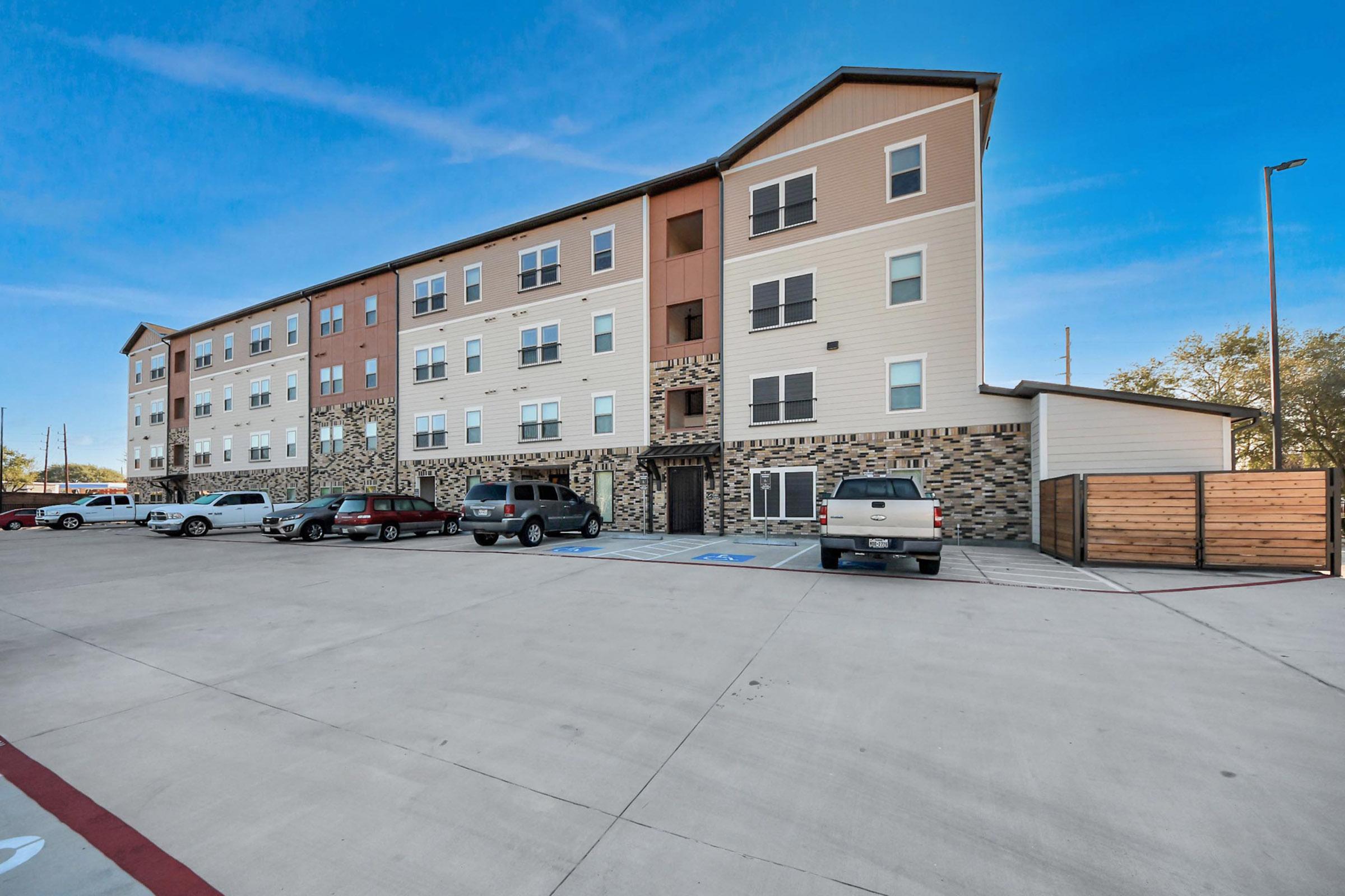 A modern multi-story apartment building with a combination of beige, orange, and stone exterior. The parking lot is visible in the foreground, featuring several parked cars. Bright blue sky in the background complements the setting.