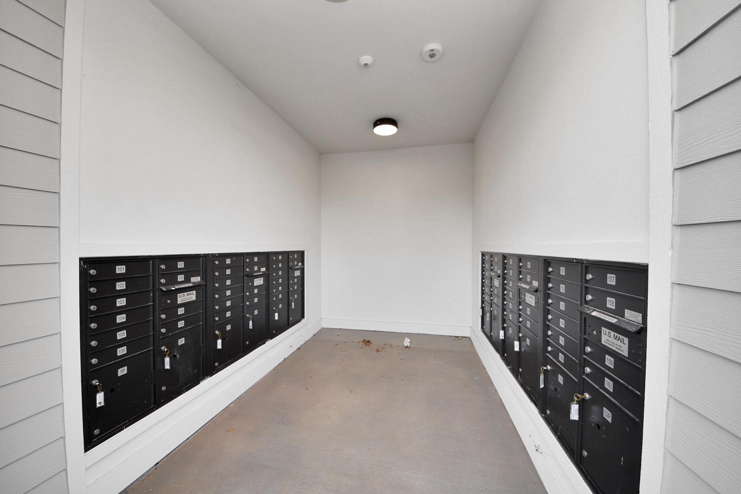 A hallway featuring row after row of black mailboxes on either side, with a light fixture on the ceiling. The space appears empty, with a clean gray floor and a few leaves scattered around. The walls are painted white, providing a bright and minimalistic atmosphere.