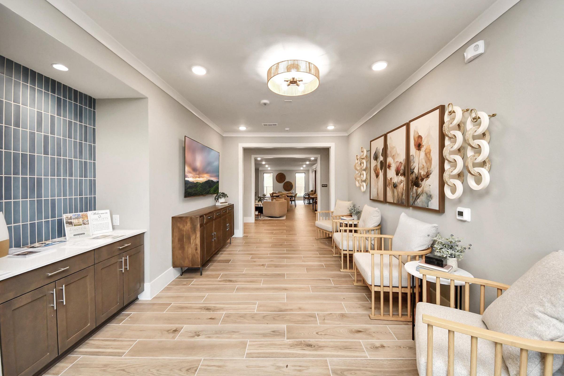 Interior view of a modern hallway featuring wooden flooring, a light-colored wall, and decorative artwork. On one side, there are stylish chairs and a wooden cabinet with a countertop. The opposite side has a blue tiled wall and leads to a spacious common area with natural light. The overall ambiance is inviting and contemporary.