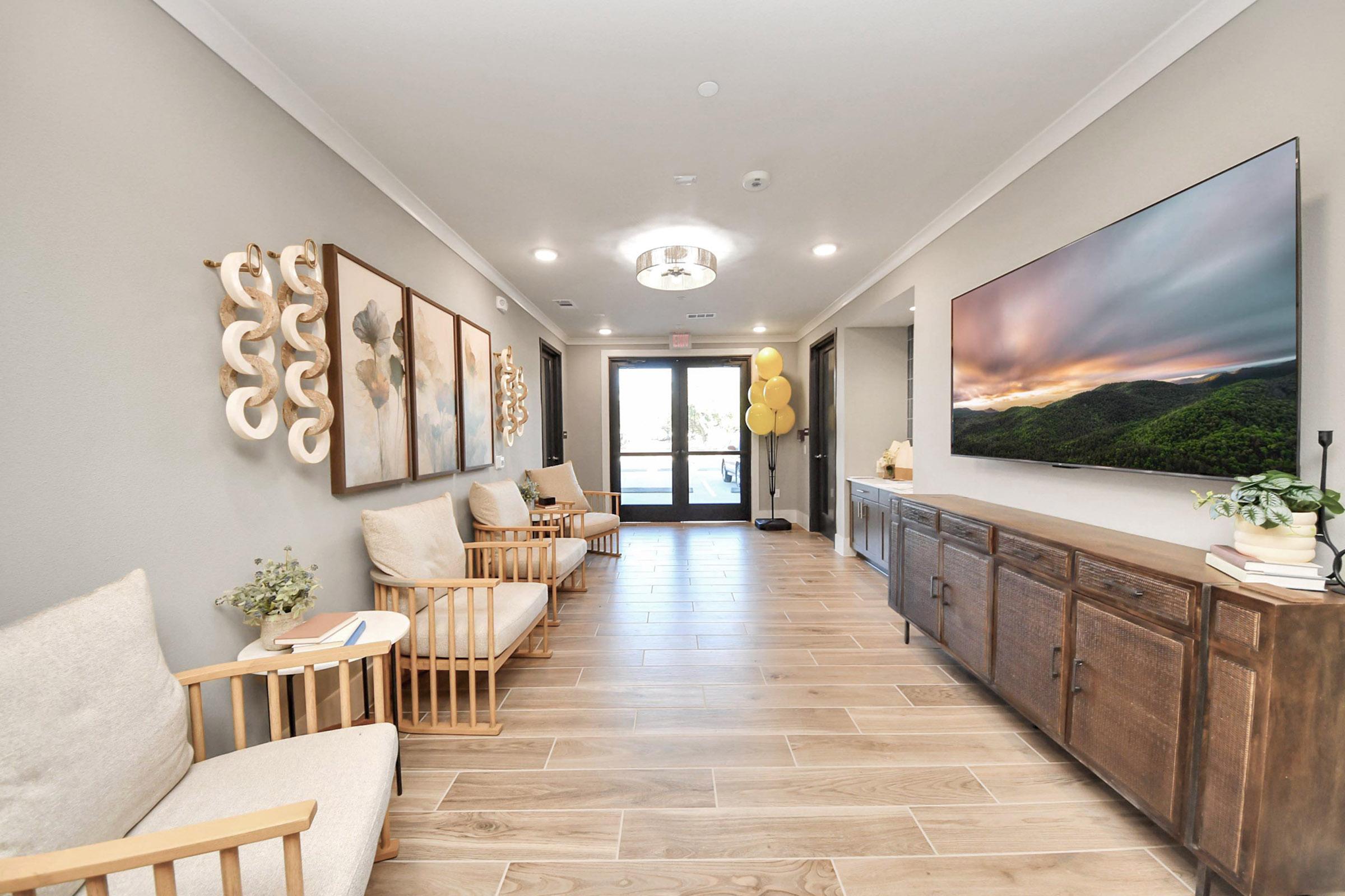 A modern hallway featuring wooden flooring, soft furniture, and decorative wall art. On the right, a large TV displays a tranquil landscape. The entrance at the end of the hallway is framed by glass doors, with yellow balloons adding a festive touch.