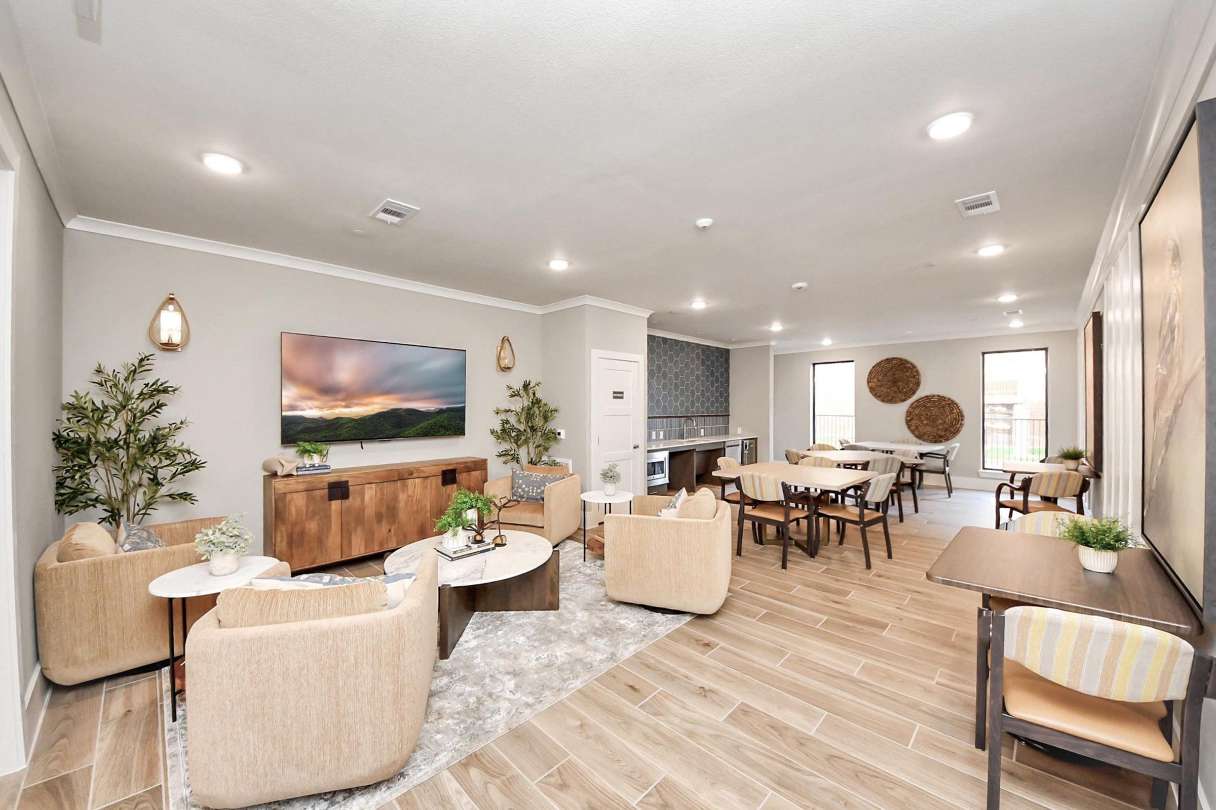 A spacious, modern lounge area featuring comfortable seating with beige chairs and a wooden coffee table, positioned around a wall-mounted TV. Light-colored walls, wood flooring, and decorative plants create a welcoming ambiance. A dining area with tables and chairs is visible in the background.