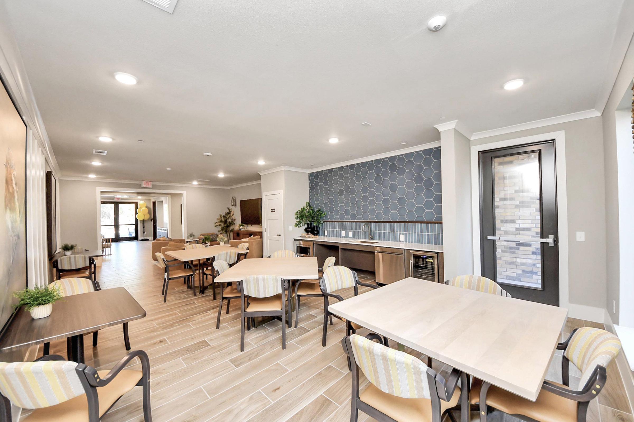A spacious dining area featuring several tables and chairs, with a modern decor. The walls are light-colored, complemented by a patterned accent wall. There are plants and a large window letting in natural light, creating a welcoming atmosphere.