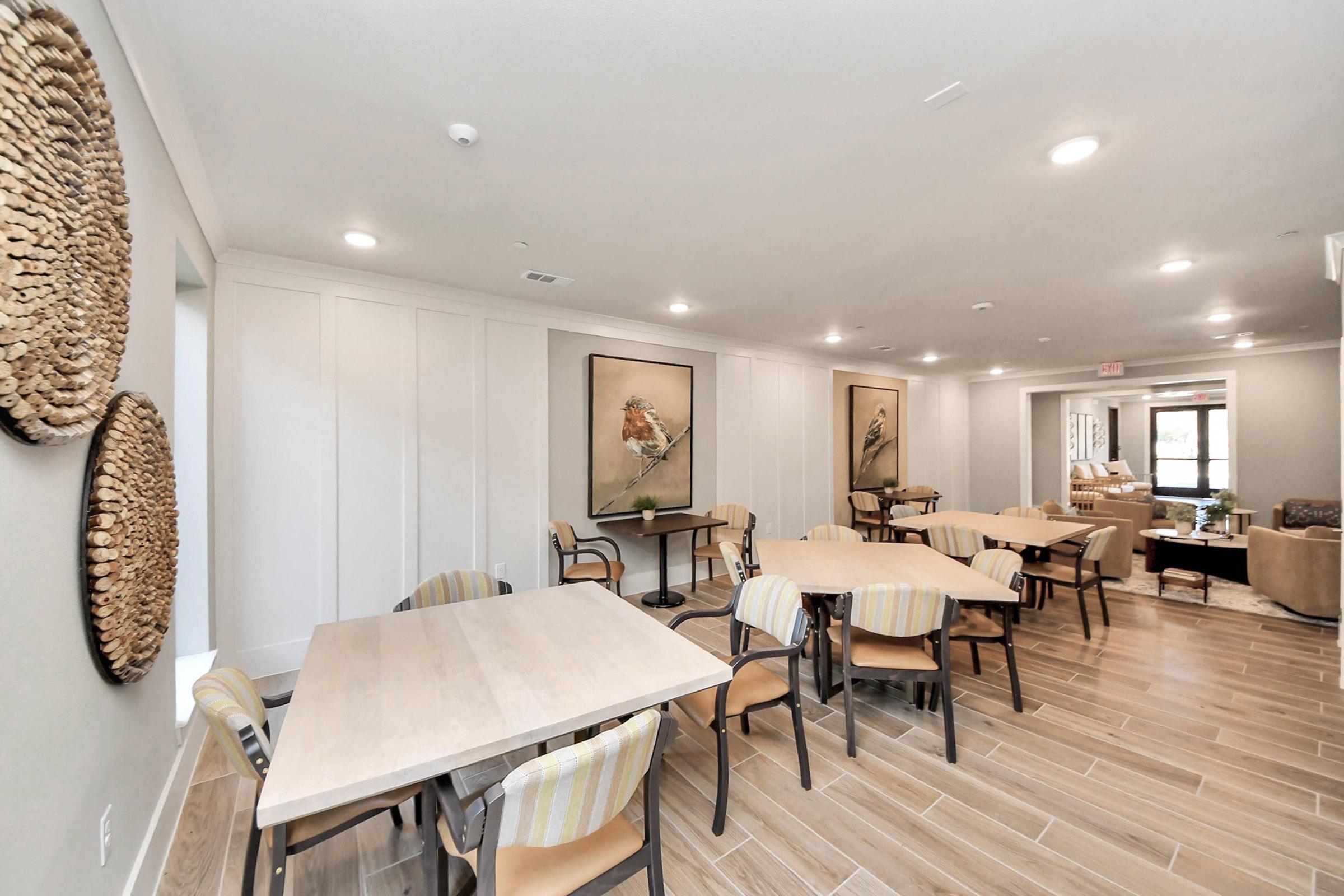 A modern dining area with wooden tables and striped chairs. There are two large framed bird artworks on the walls. Natural light fills the space, and decorative woven wall hangings add texture. The flooring is made of light-colored tiles, creating a warm and inviting atmosphere.