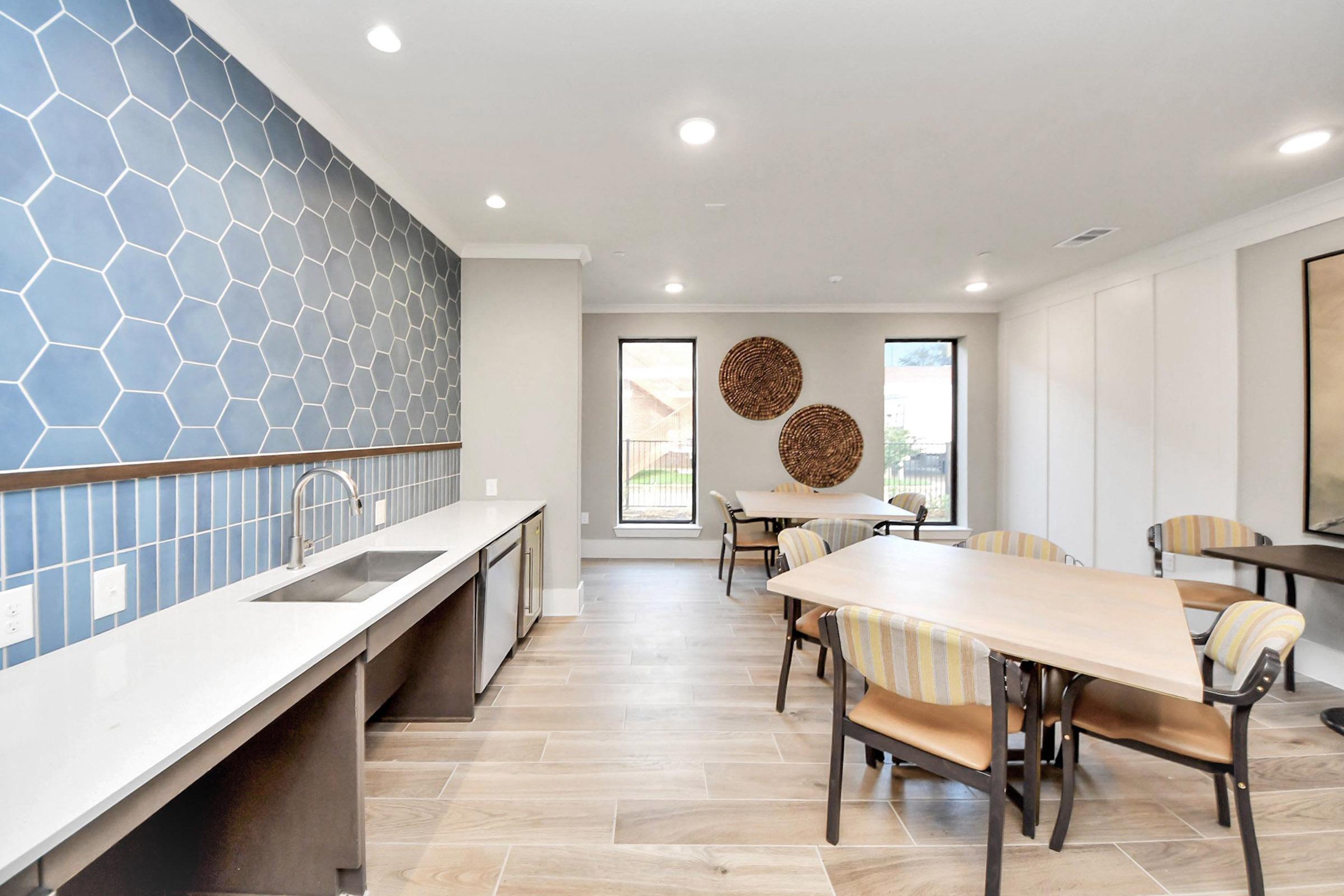 Modern dining area featuring a honeycomb-patterned blue accent wall, white countertops, and wooden flooring. The space includes tables and comfortable chairs, with large windows allowing natural light. Decorative round wall pieces add an artistic touch to the room.