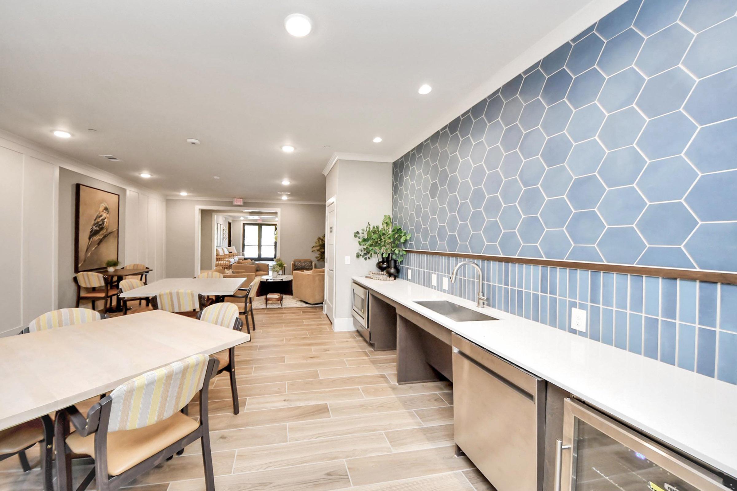 A modern kitchen with a hexagonal blue tile accent wall, white countertops, and stainless steel appliances. There are wooden tables and chairs arranged, creating a welcoming dining area. A potted plant adds a touch of greenery, and a living room can be seen in the background.