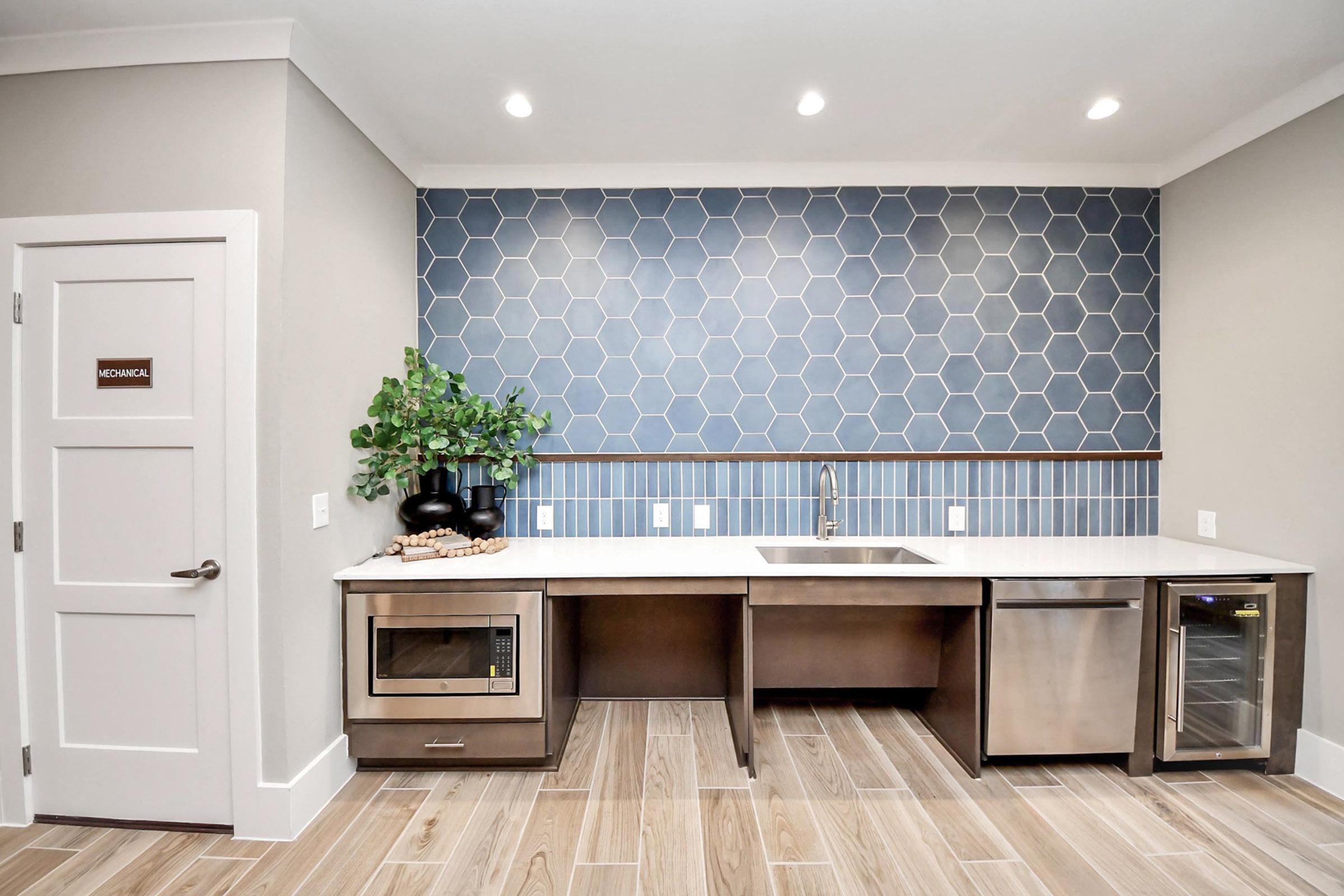 A modern kitchen setup featuring a blue hexagonal tiled wall, a stainless steel sink, a built-in microwave, and a wine cooler. The counter has decorative items and a green plant. There’s a white door labeled "Mechanical" on the left. The flooring consists of light brown tiles.