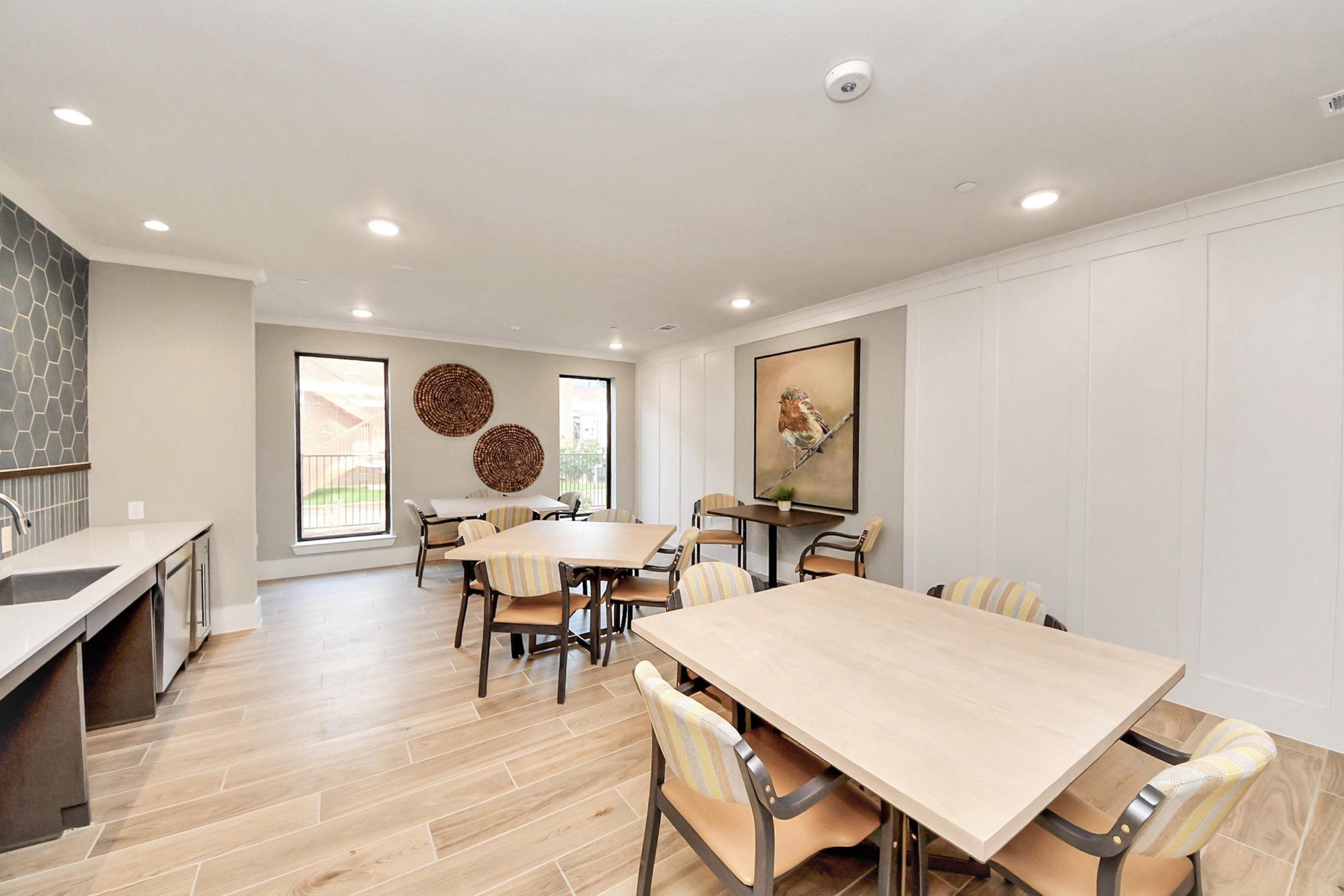 Interior view of a modern communal space featuring multiple round tables with chairs, large windows providing natural light, a sleek kitchen area, and decorative wall art, creating a welcoming atmosphere for gatherings or meetings.