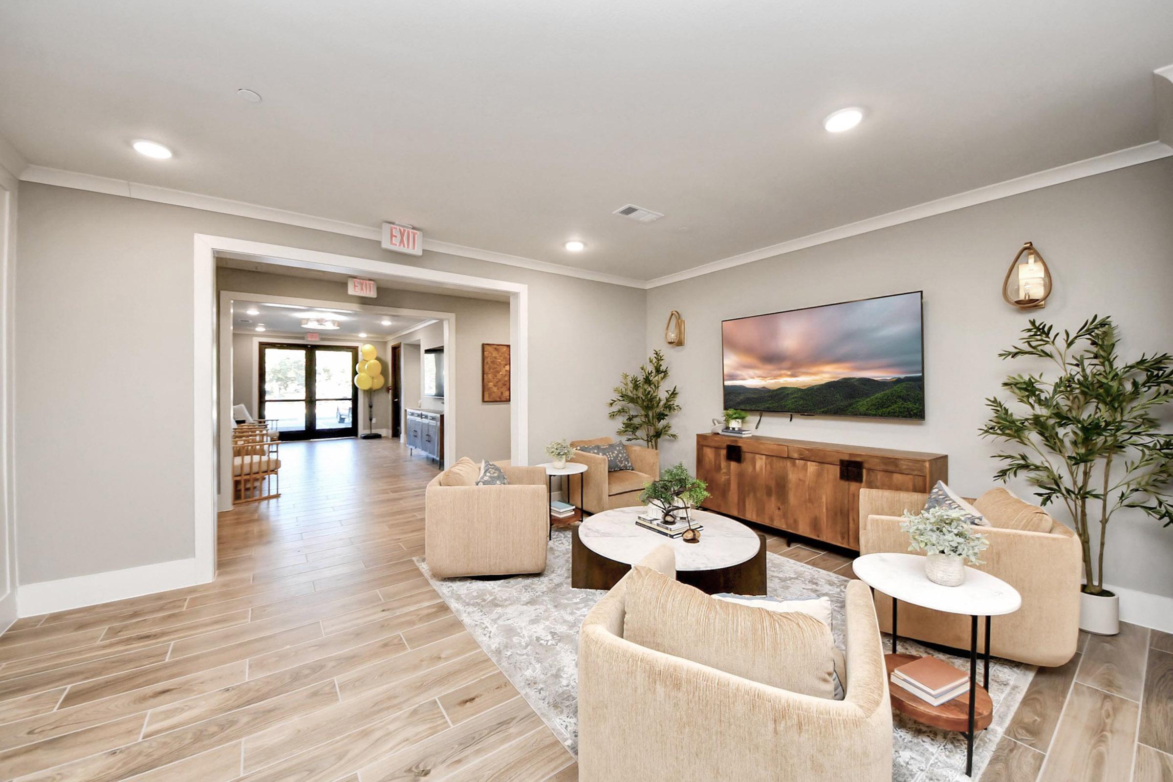 A modern lounge area featuring two beige armchairs, a round coffee table, and decorative plants. A large TV is mounted on the wall above a wooden console. The space has light-colored walls and flooring, with an entrance leading to another room visible in the background. The atmosphere is inviting and contemporary.