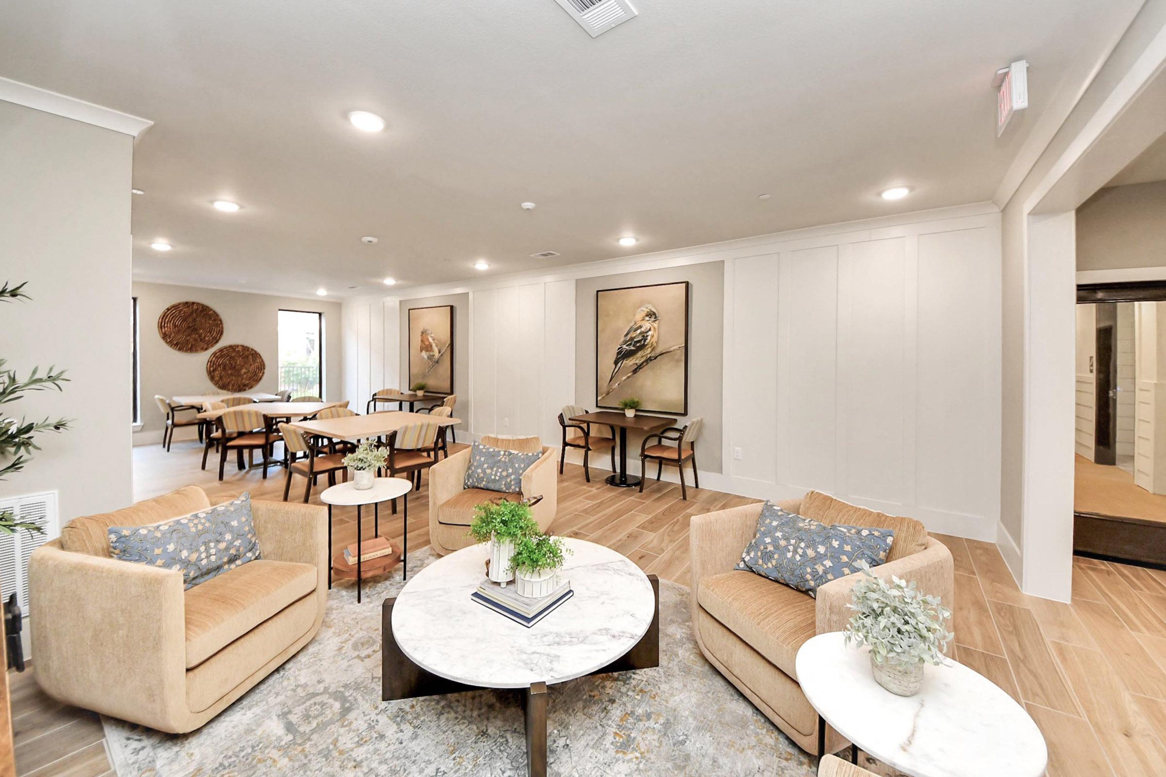 A modern indoor lounge area featuring comfortable seating with beige armchairs and a round marble coffee table. The walls are adorned with framed artwork of birds, and circular decorative pieces. Bright lighting enhances the open space, which includes dining tables and chairs in a cozy, welcoming design.