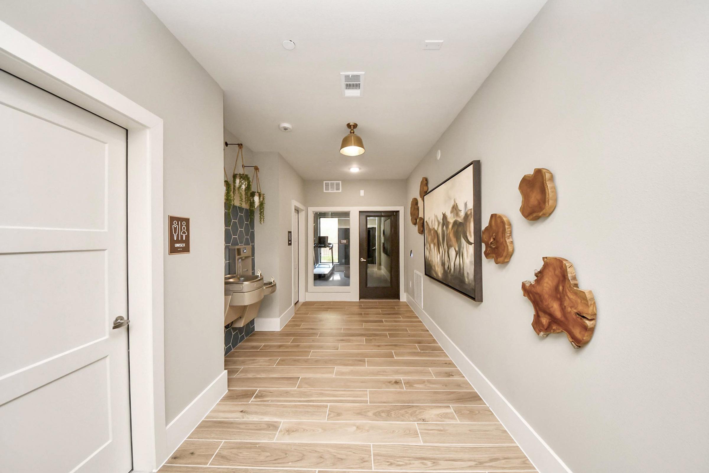A modern hallway featuring light gray walls and wooden flooring, with a closed white door on the left. There is a contemporary art piece on the right wall and a water fountain nearby. The hallway leads to a glass door, providing access to an outdoor area, with decorative elements adding warmth to the space.