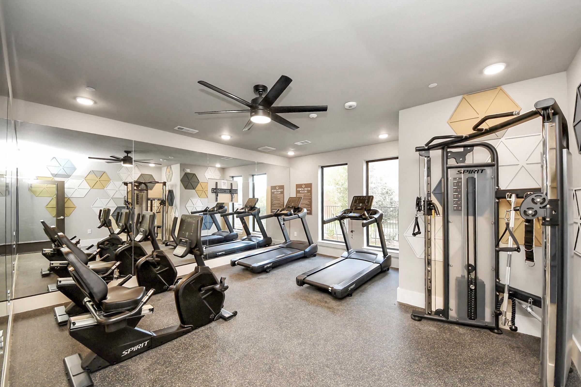 A modern gym interior featuring several treadmills, a stationary bike, and a multi-gym strength training machine. The space has large windows allowing natural light, and the walls are adorned with geometric patterns. The flooring is a dark, textured surface, creating a sleek, contemporary atmosphere.
