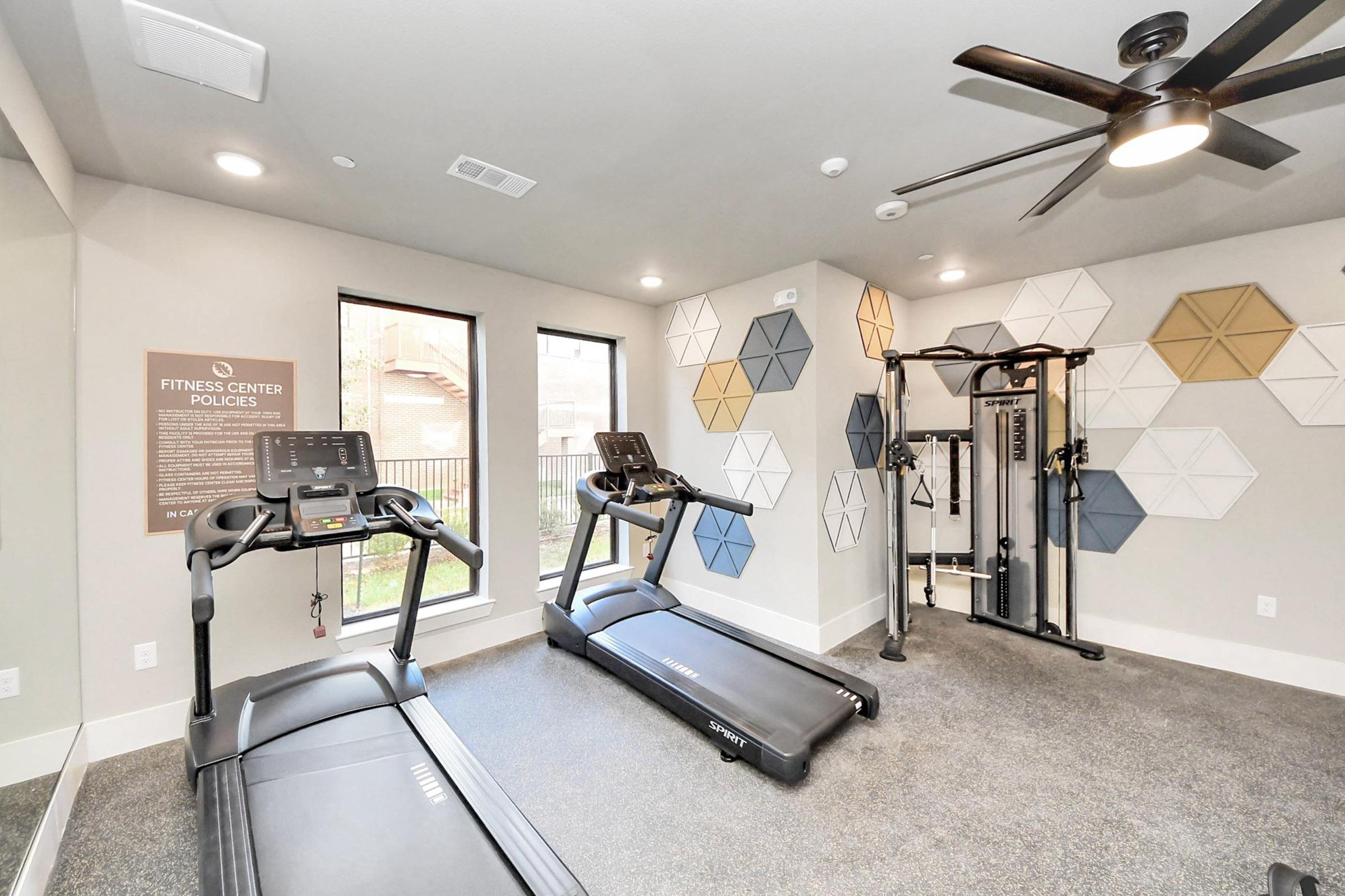 A well-lit fitness center featuring two treadmills, a weight training machine, and a wall decorated with geometric patterns in neutral tones. Large windows provide natural light, enhancing the modern and inviting atmosphere of the workout space.