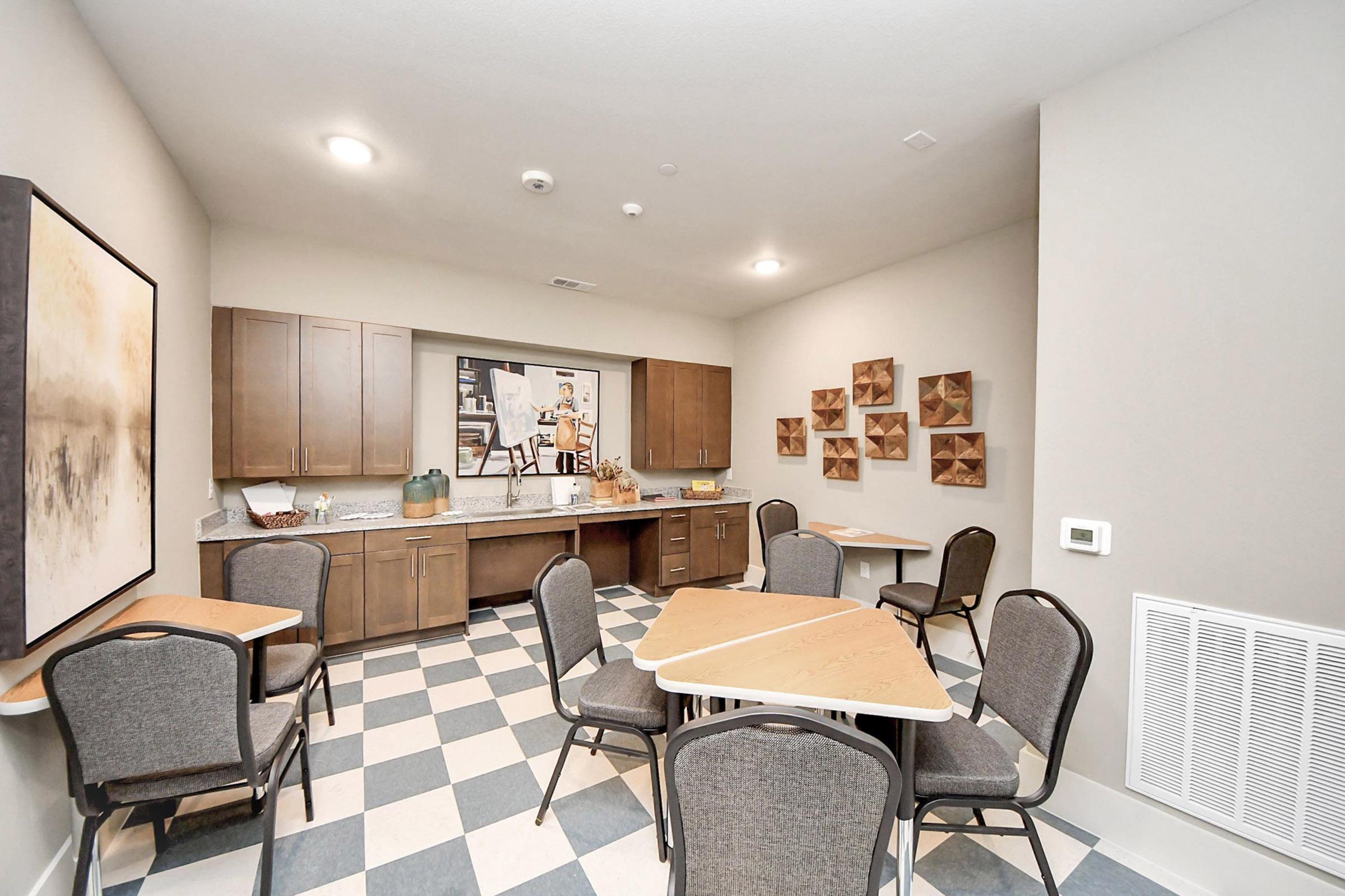 Interior view of a small communal dining area featuring four tables with chairs, a kitchenette with wooden cabinets, and decorative wall art. The floor has a checkered pattern, and the space is well-lit with soft ceiling lights, creating a cozy atmosphere.