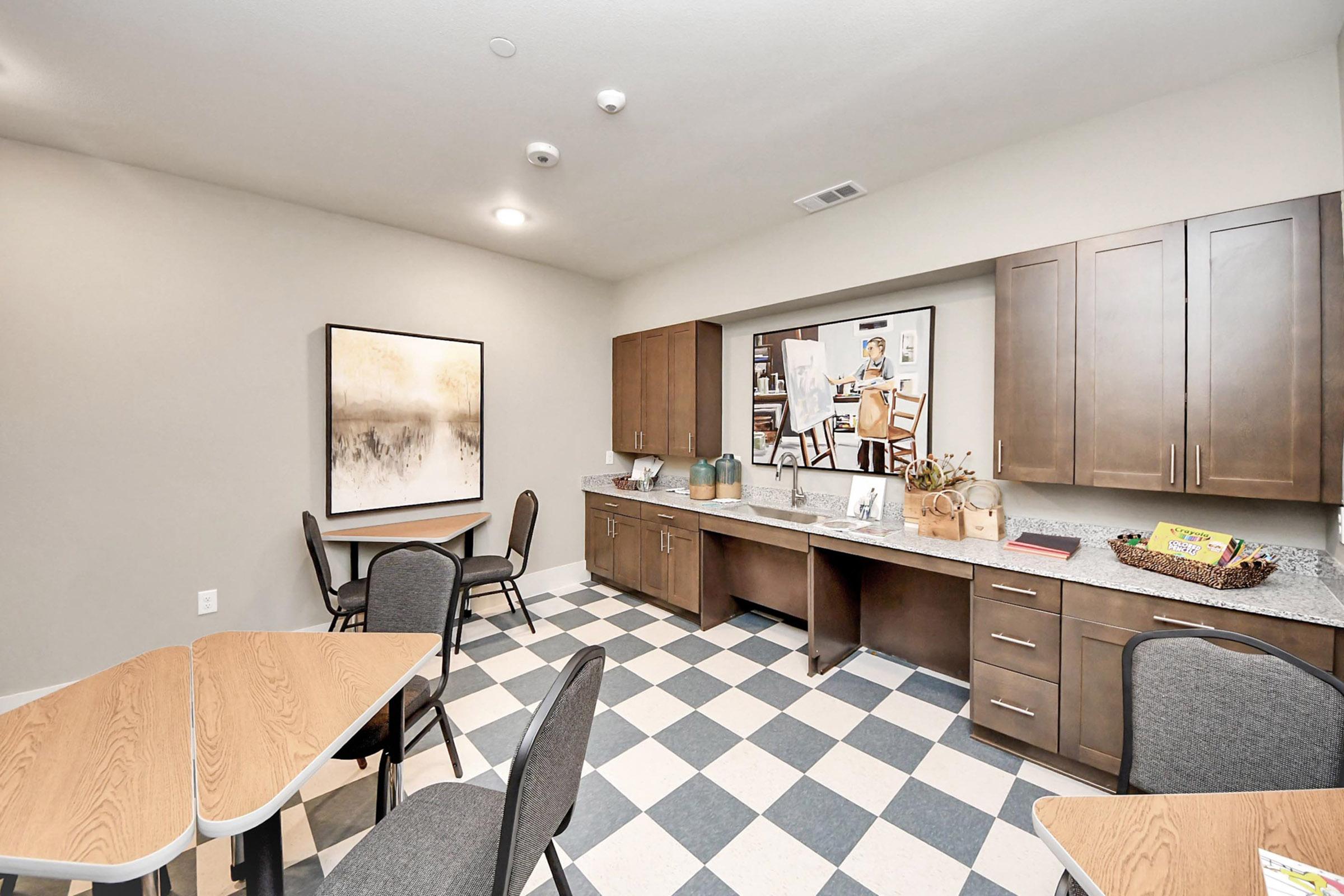 A modern, well-lit community room featuring gray walls, a large abstract painting, and a kitchenette with dark wood cabinets. There are multiple tables and chairs in a checkered floor pattern, creating a welcoming space for gatherings or meetings.