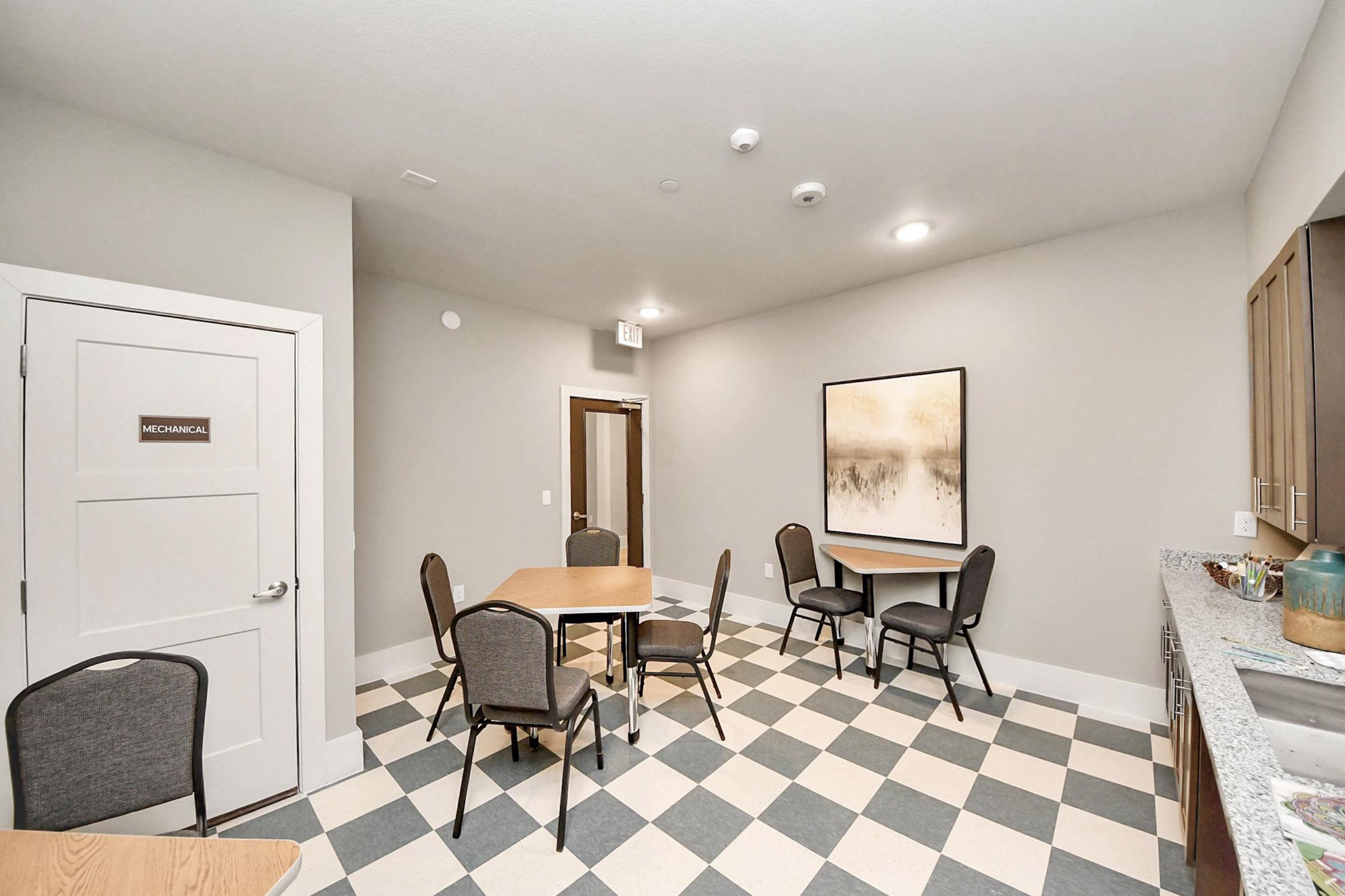 A well-lit room with a checkered floor, featuring a table with four chairs, a kitchenette area, and a decorative wall art piece. The door on the left is labeled "MECHANICAL." The walls are painted in a neutral tone, creating a calm and inviting atmosphere.