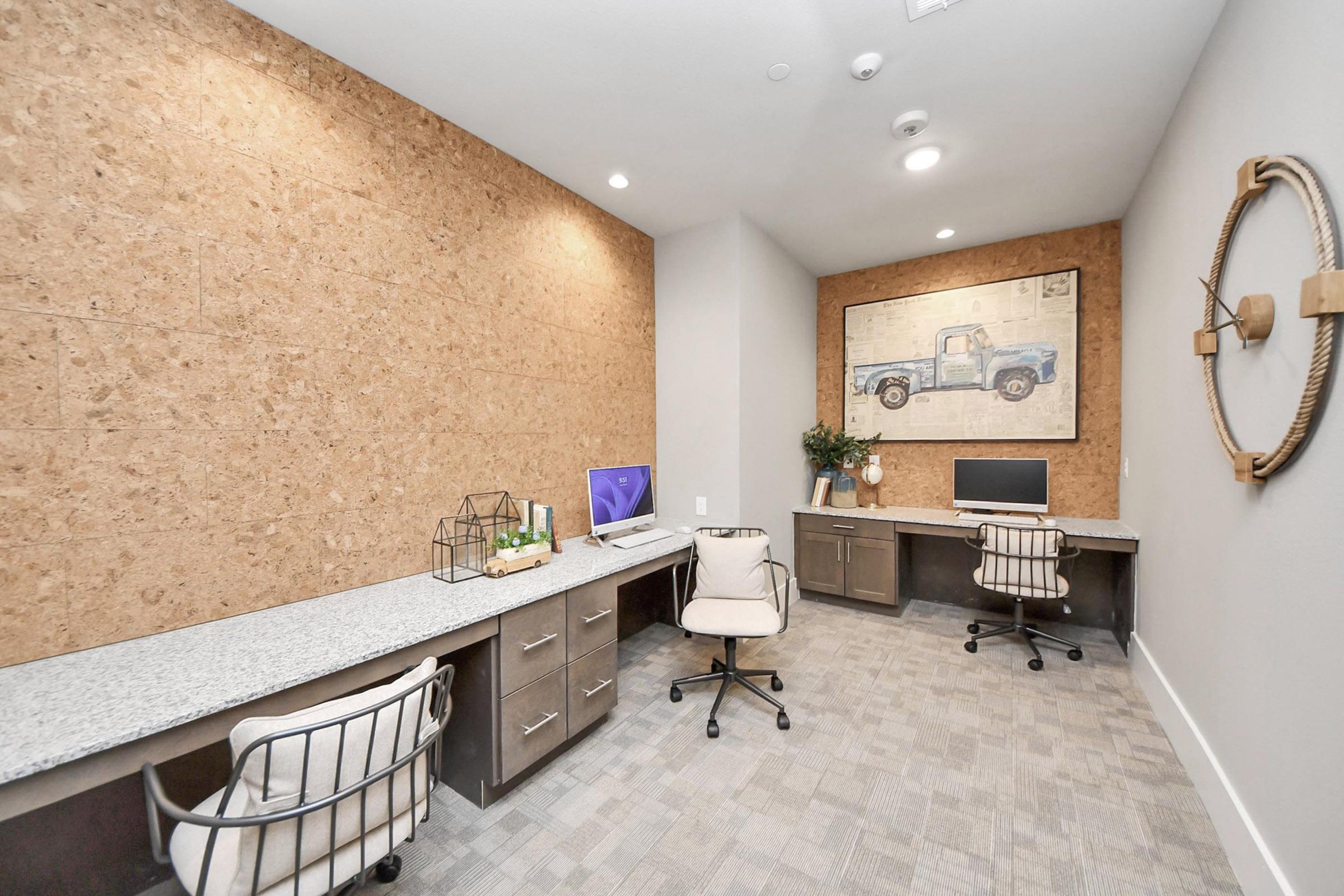 A modern office space featuring two desks with comfortable chairs, grey countertops, and a cork wall. One desk has a computer setup, while the other is adorned with decorative items. A vintage truck print is mounted on the wall, adding a touch of character to the room.
