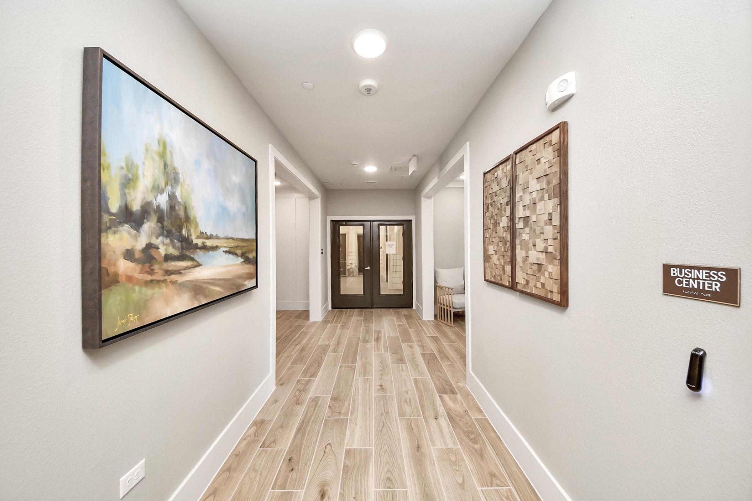 A modern hallway with light gray walls and wooden tile flooring. On the left, there is a large landscape painting with earthy tones. On the right, a textured wall art piece made of natural materials. A sign for a business center is displayed on the wall near a door at the end of the hallway.