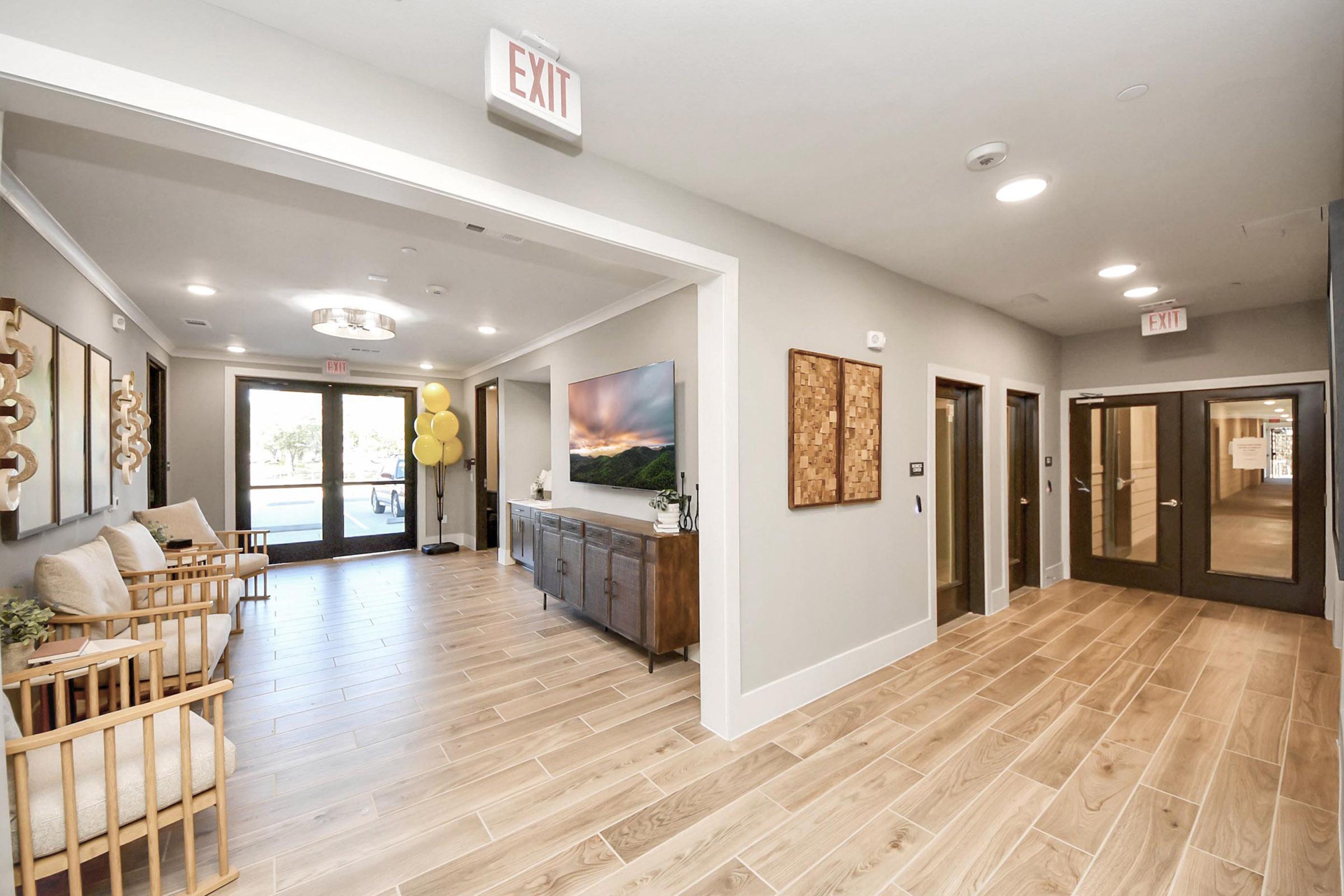 A well-lit, modern lobby area featuring wooden flooring, a seating arrangement with light-colored chairs, and decorative wall art. The space includes a side console table, a large TV on the wall, and glass doors leading to an outdoor area. Yellow balloons add a festive touch, with visible exit signs.