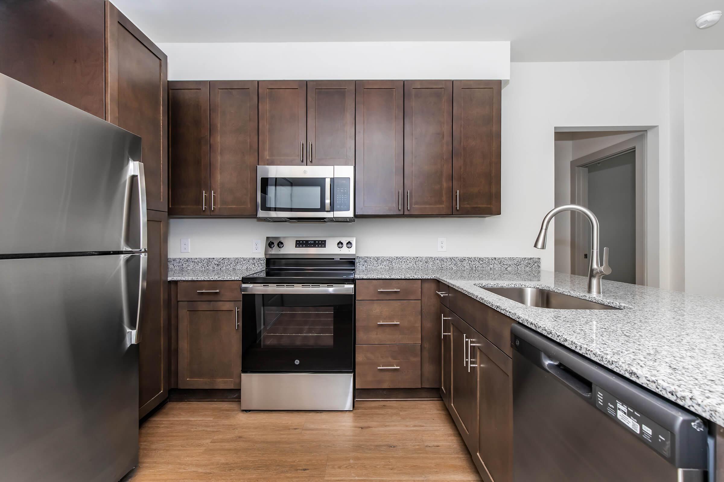 Modern kitchen featuring dark wooden cabinetry, stainless steel appliances including a refrigerator, microwave, and oven, a granite countertop, and a sink with a sleek faucet. The flooring is light wood, providing a warm contrast to the cabinetry and modern appliances.