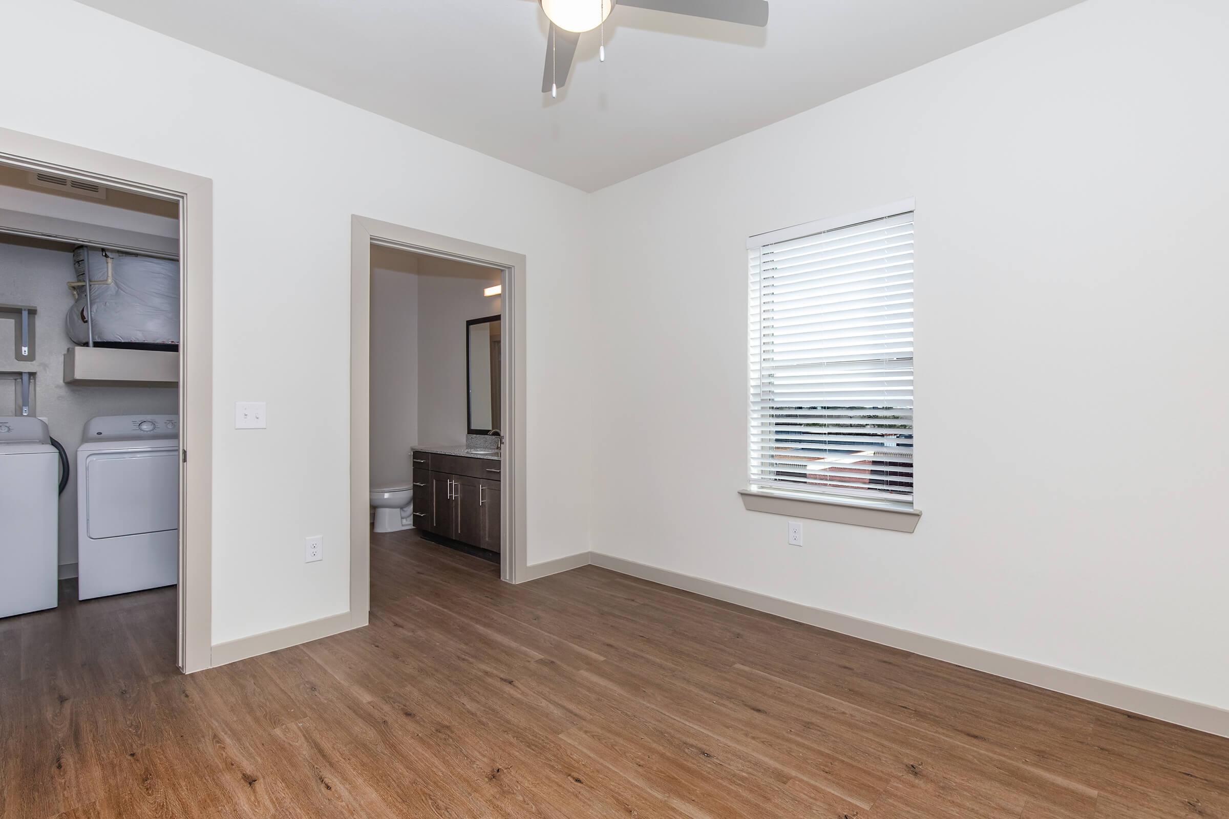 Interior view of a modern apartment featuring a spacious room with light-colored walls, a ceiling fan, and laminate flooring. To the left, there is a doorway leading to a laundry area with a washer and dryer. A bathroom is visible in the background, and a window with blinds allows natural light into the space.