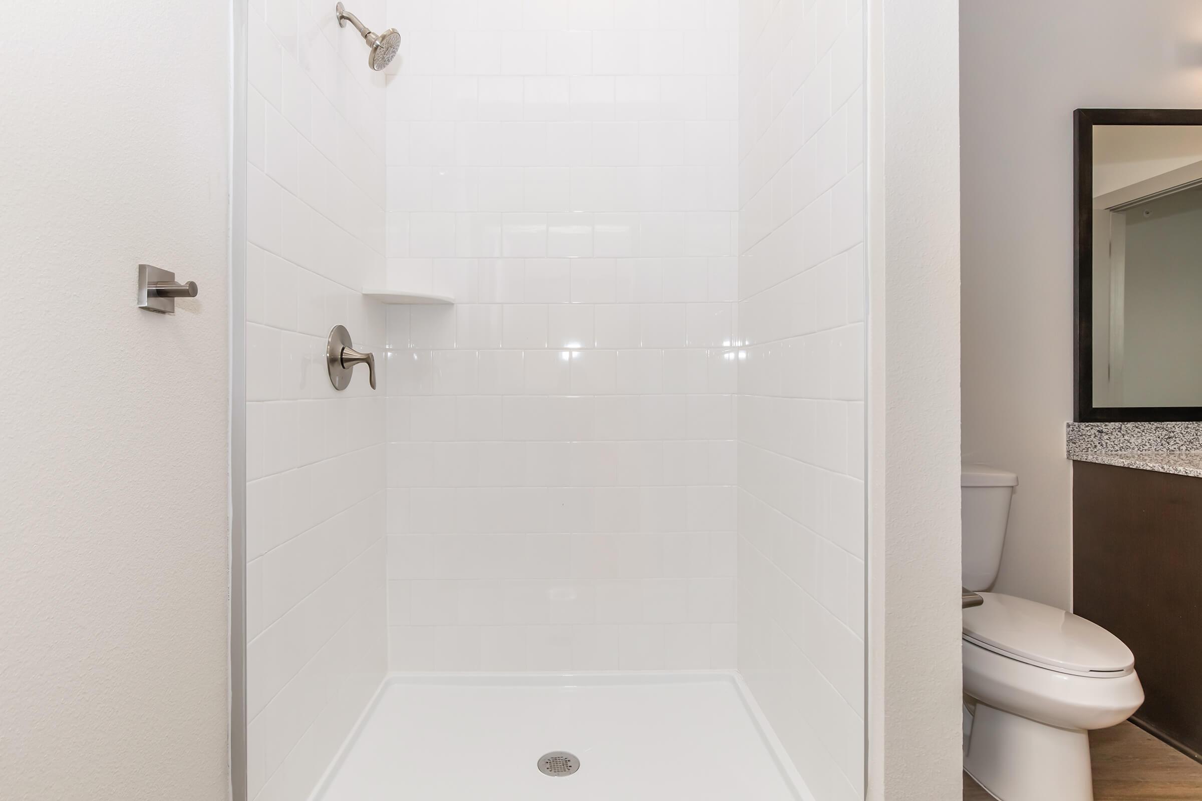 A clean, modern shower stall with white tiled walls and a stainless steel showerhead. The shower includes a small shelf for toiletries and is adjacent to a bathroom with a toilet and a dark, granite countertop. The space is bright and uncluttered.