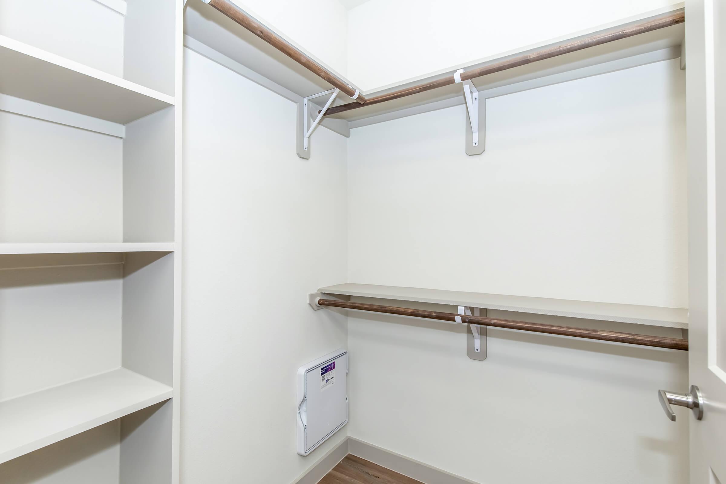 An empty closet featuring white walls, a shelf and hanging rods made of wood, and some built-in shelving on the left. The floor is a light-colored wood, and a small utility panel is visible near the floor. The overall decor is simple and functional, suitable for storage.