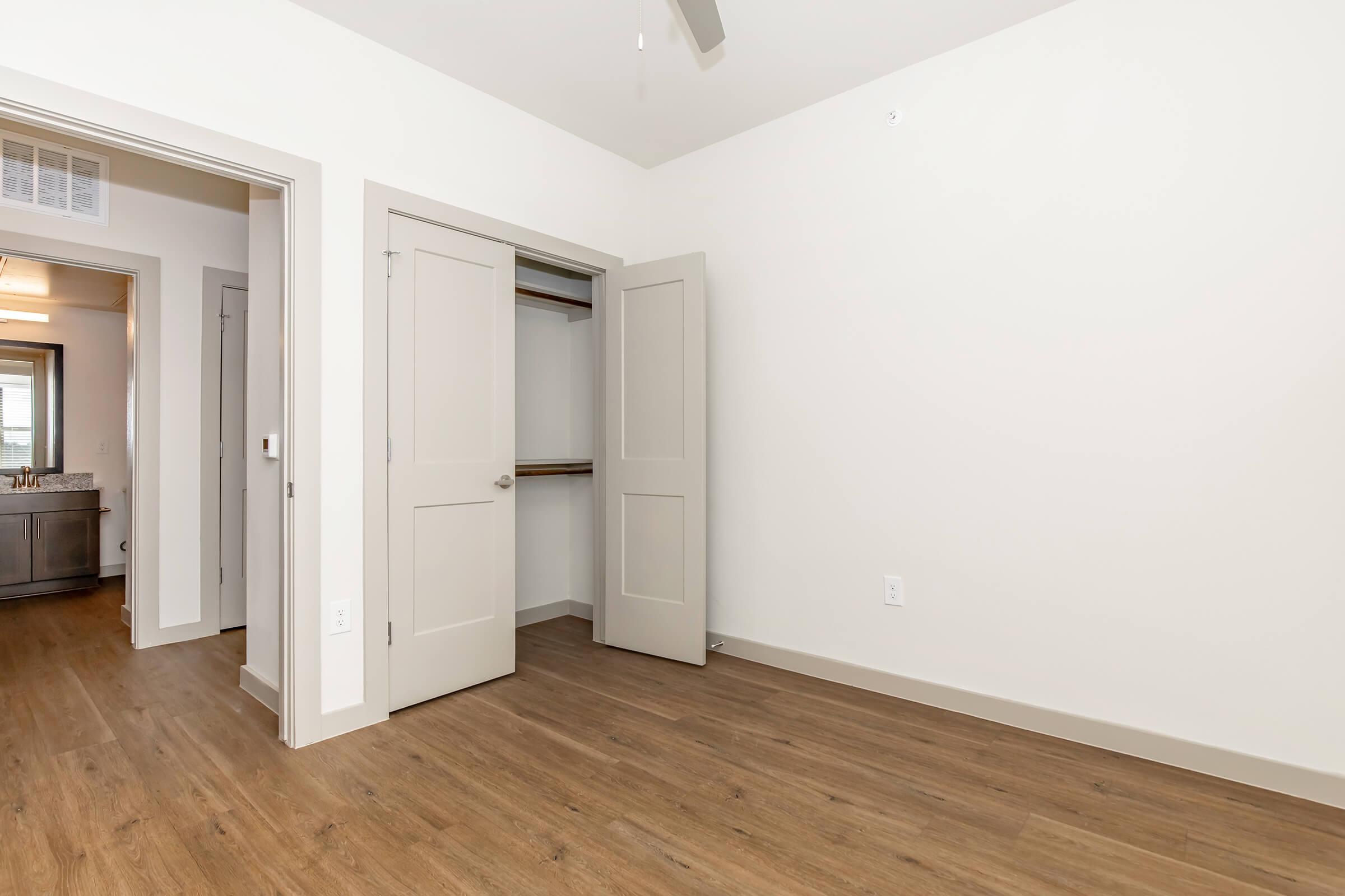 Empty interior of a room featuring a light-colored wall, a brown laminate floor, and an open closet with sliding doors. A doorway leads to a bathroom area with visible fixtures. The room is well-lit and spacious, showcasing a minimalist design.