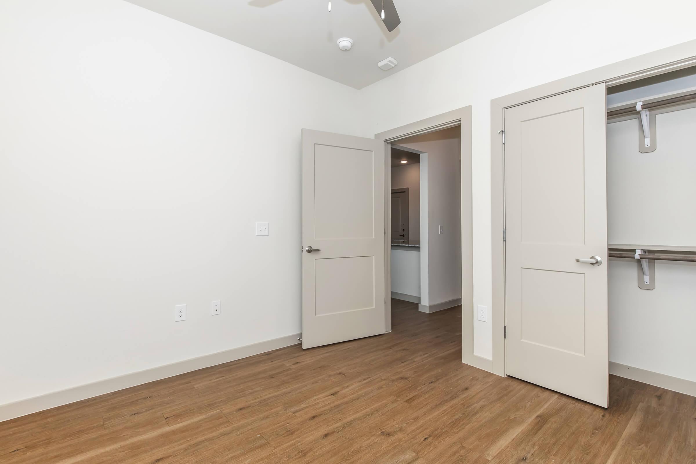 A spacious, empty room featuring light-colored walls and wood-like flooring. Two beige doors are open, leading to a closet and a hallway. A ceiling fan is visible, and a doorway in the background suggests additional space or rooms. The overall atmosphere is bright and modern.