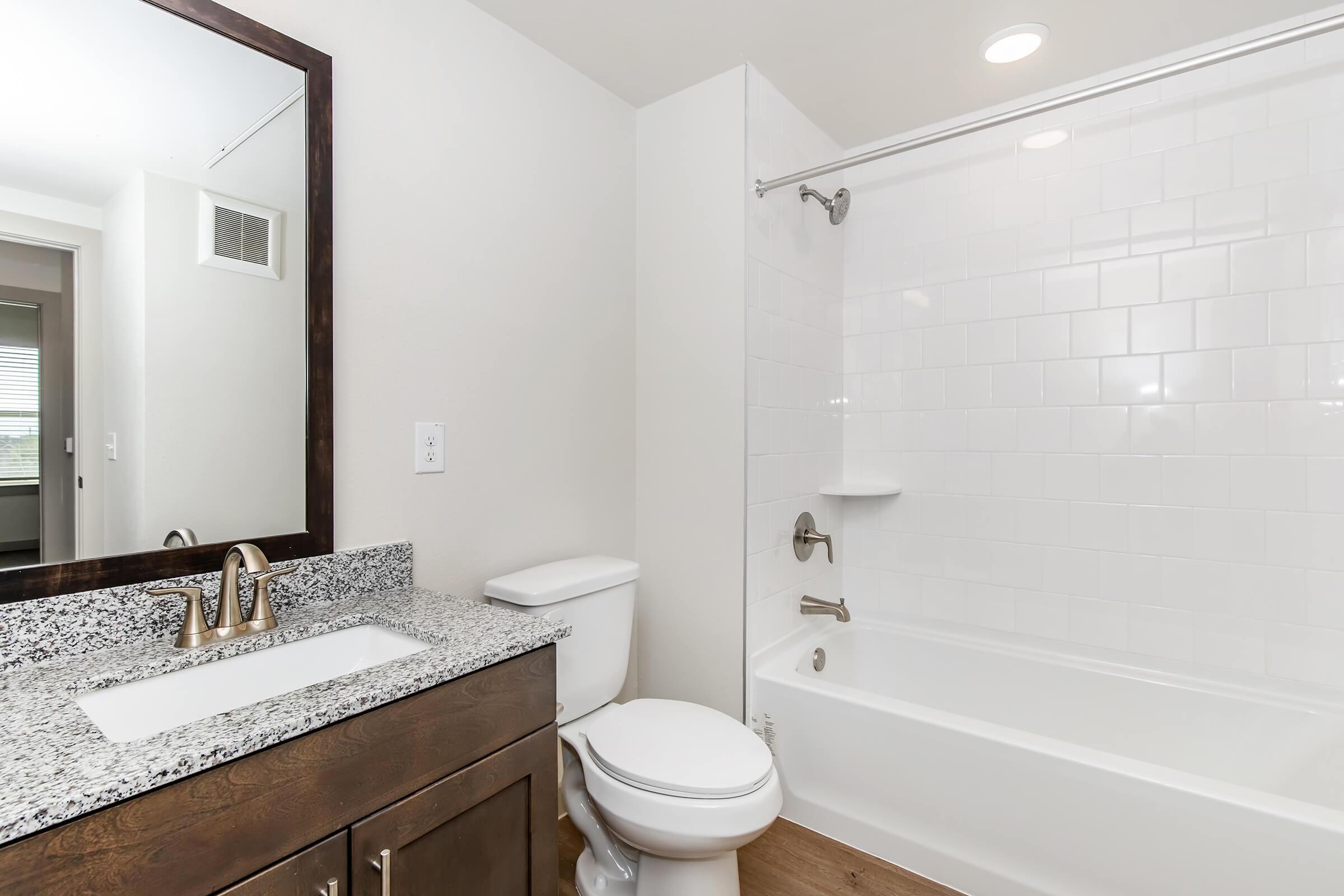 A modern bathroom featuring a white tiled shower/tub combination, a granite countertop sink with a mirror above, and a clean, minimalist design. The decor includes light-colored walls and wooden flooring for a warm and inviting atmosphere.