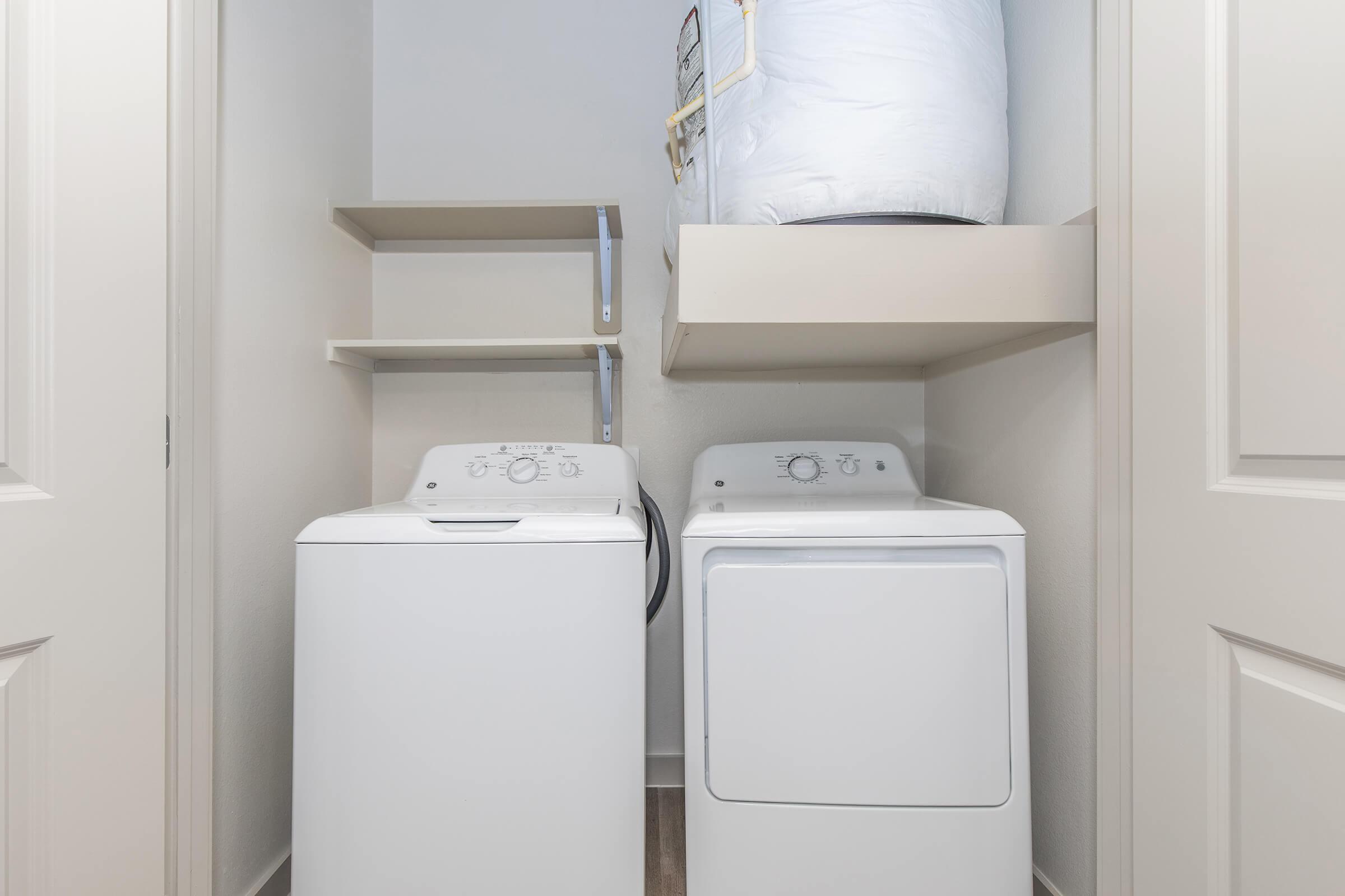 A laundry closet featuring a white washing machine and a white dryer side by side. Above them, there is a shelf with a decorative storage bin. The walls are painted in a light color, creating a bright and clean atmosphere. Doors are partially closed, adding a tidy appearance to the space.