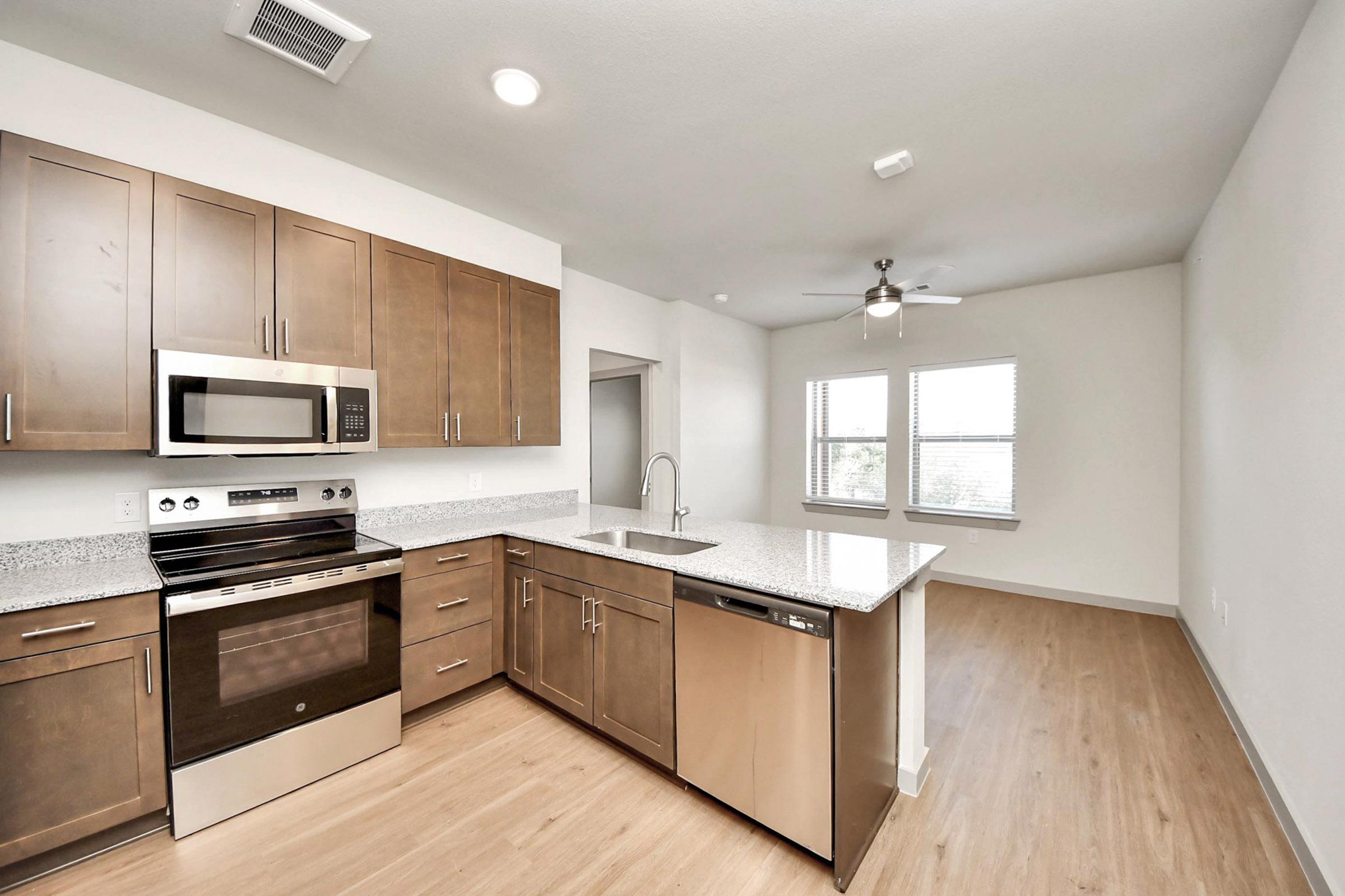 Modern kitchen featuring brown cabinetry, stainless steel appliances including an oven, microwave, and dishwasher, with a granite countertop. A ceiling fan is visible, and large windows provide natural light. The space has a light wood floor and an open layout leading to a living area.