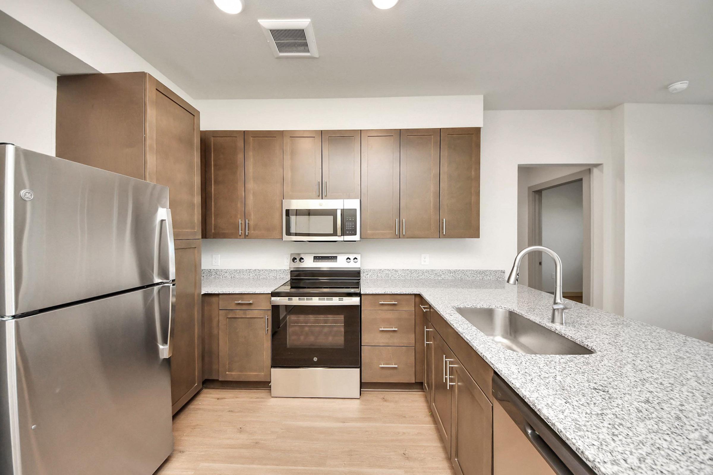 A modern kitchen featuring stainless steel appliances, including a refrigerator, microwave, and oven. The cabinetry is light brown, complemented by a granite countertop. The kitchen has a spacious layout with hardwood flooring and a sink with a stylish faucet.