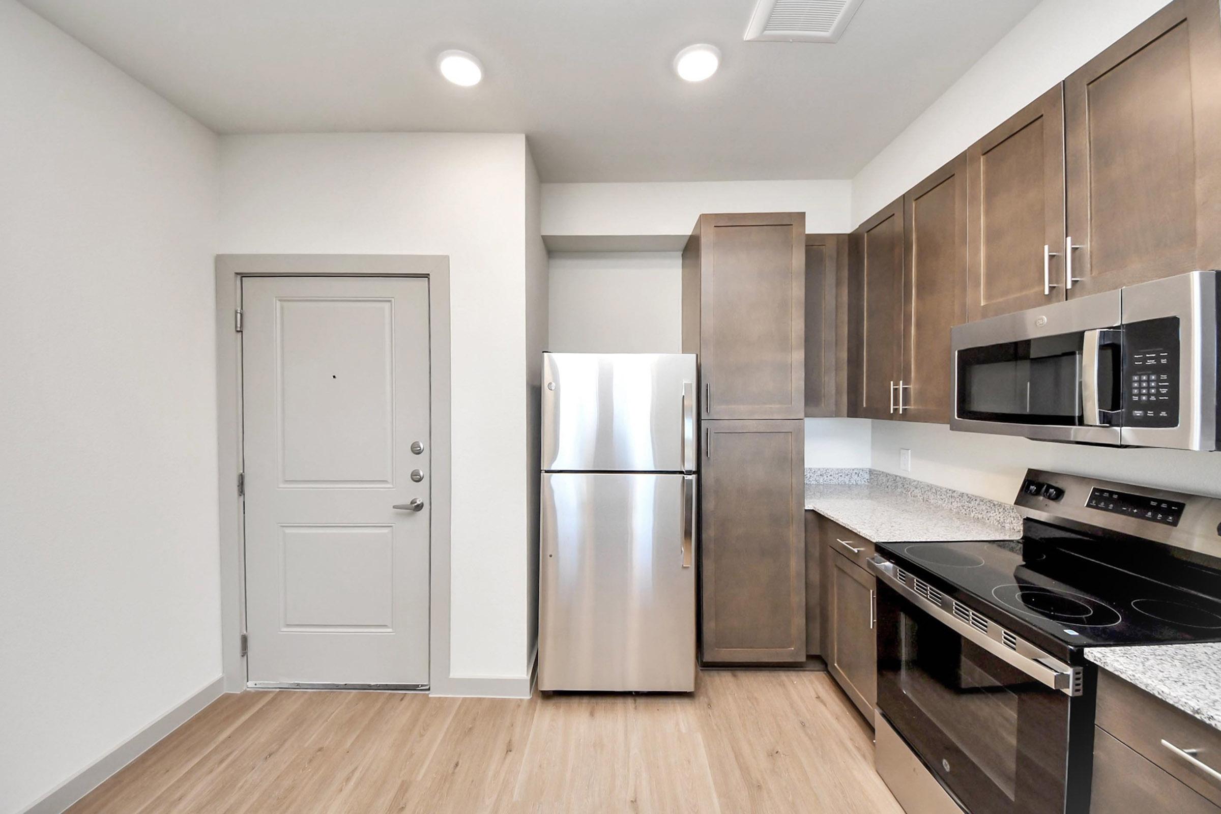 Modern kitchen featuring stainless steel appliances, including a refrigerator and microwave, with dark wood cabinetry and granite countertops. The space is well-lit with overhead lighting and has a door leading outside. The flooring is a light wood tone, creating a cozy atmosphere.