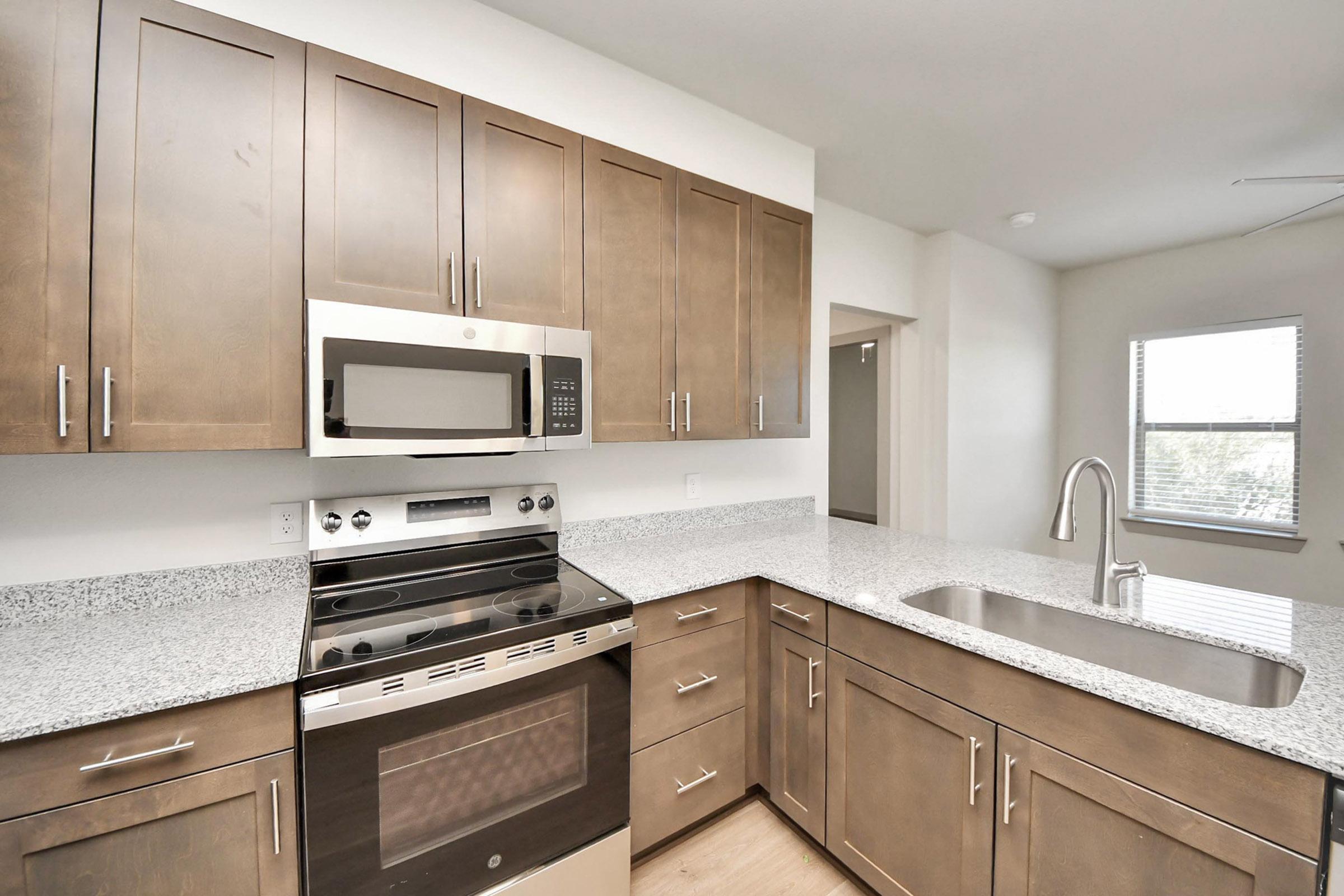 Modern kitchen featuring dark wood cabinetry, stainless steel appliances including a microwave and oven, and a spacious granite countertop with a sink. Natural light enters through a nearby window, highlighting the contemporary design and open layout.